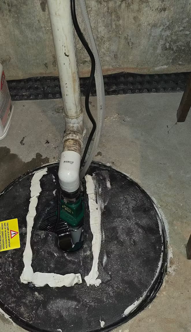 Black sump lid with the pump visible through a caulked cutout, white PVC discharge pipe and a clear hose rising from it