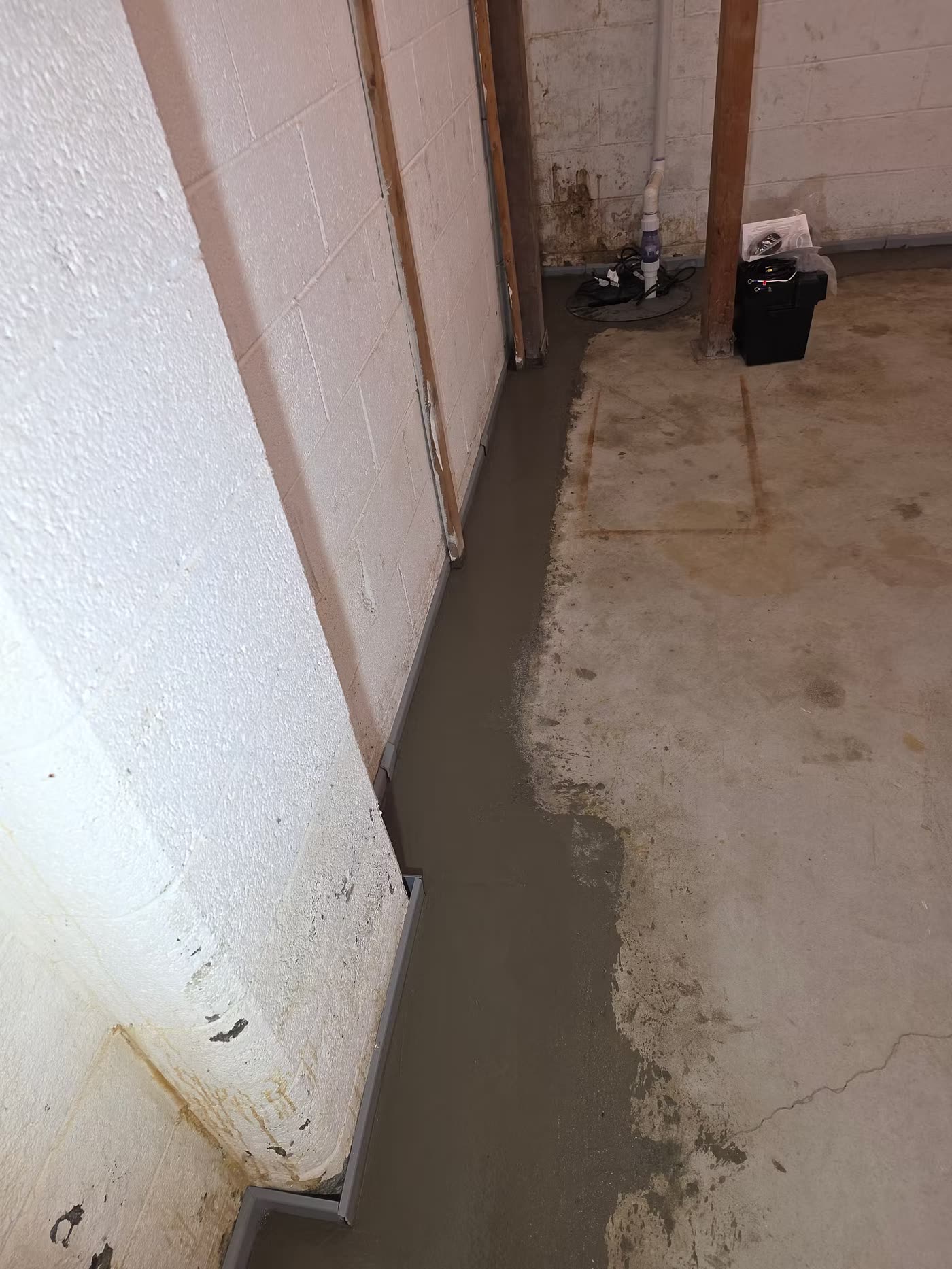 Interior French drain along the base of a painted block wall, fresh concrete beside it, sump lid and discharge pipe in the corner