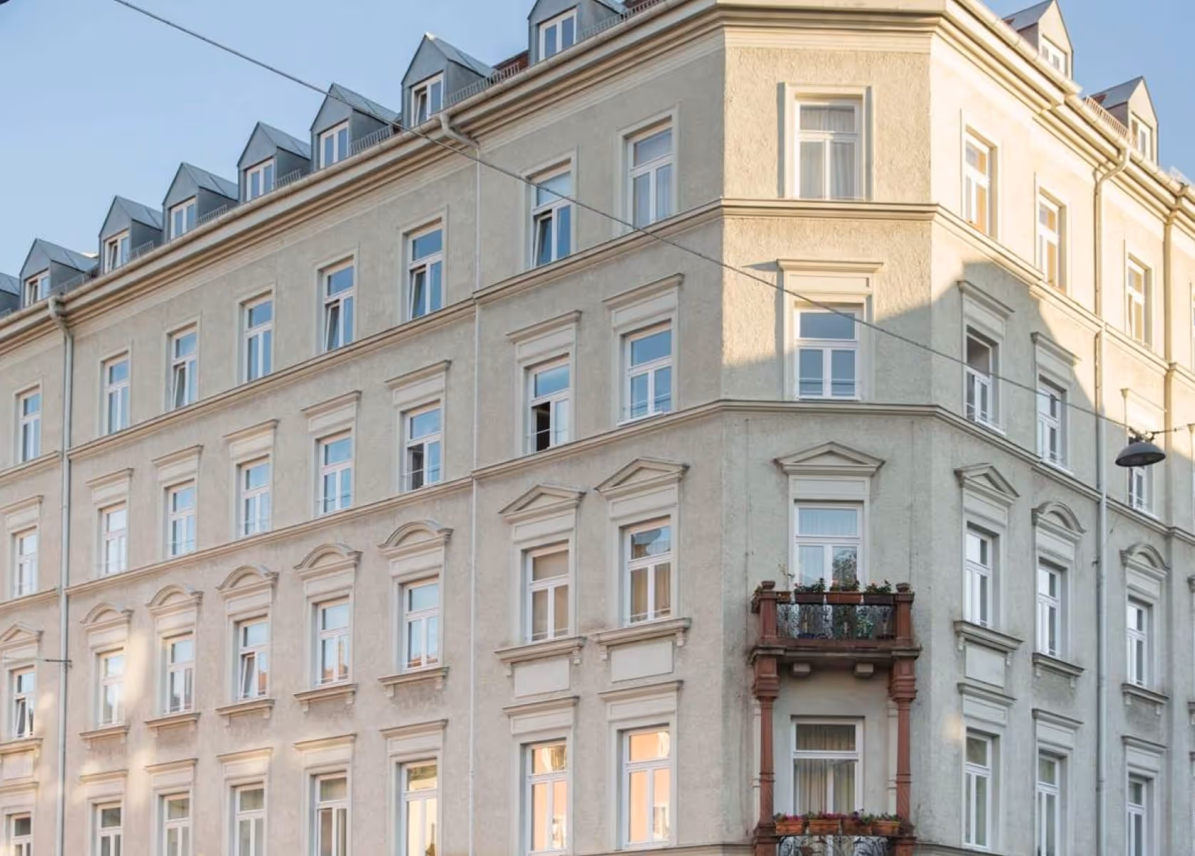 Helles mehrstöckiges Wohngebäude mit klassischer Fassade, vielen Fenstern und einem kleinen Balkon, aufgenommen an einer Straßenecke bei Tageslicht.