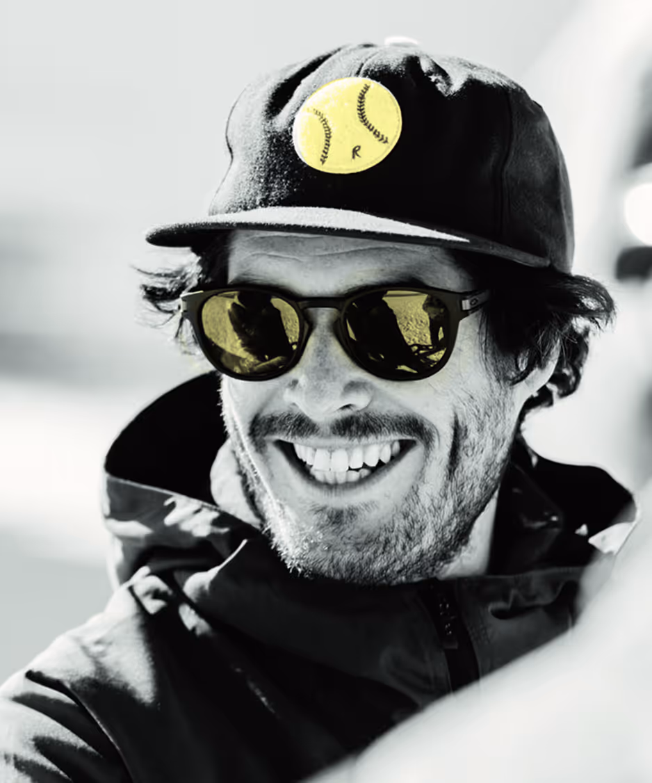 Homme souriant portant une casquette noire avec un logo jaune de balle de tennis et des lunettes de soleil à verres jaunes.