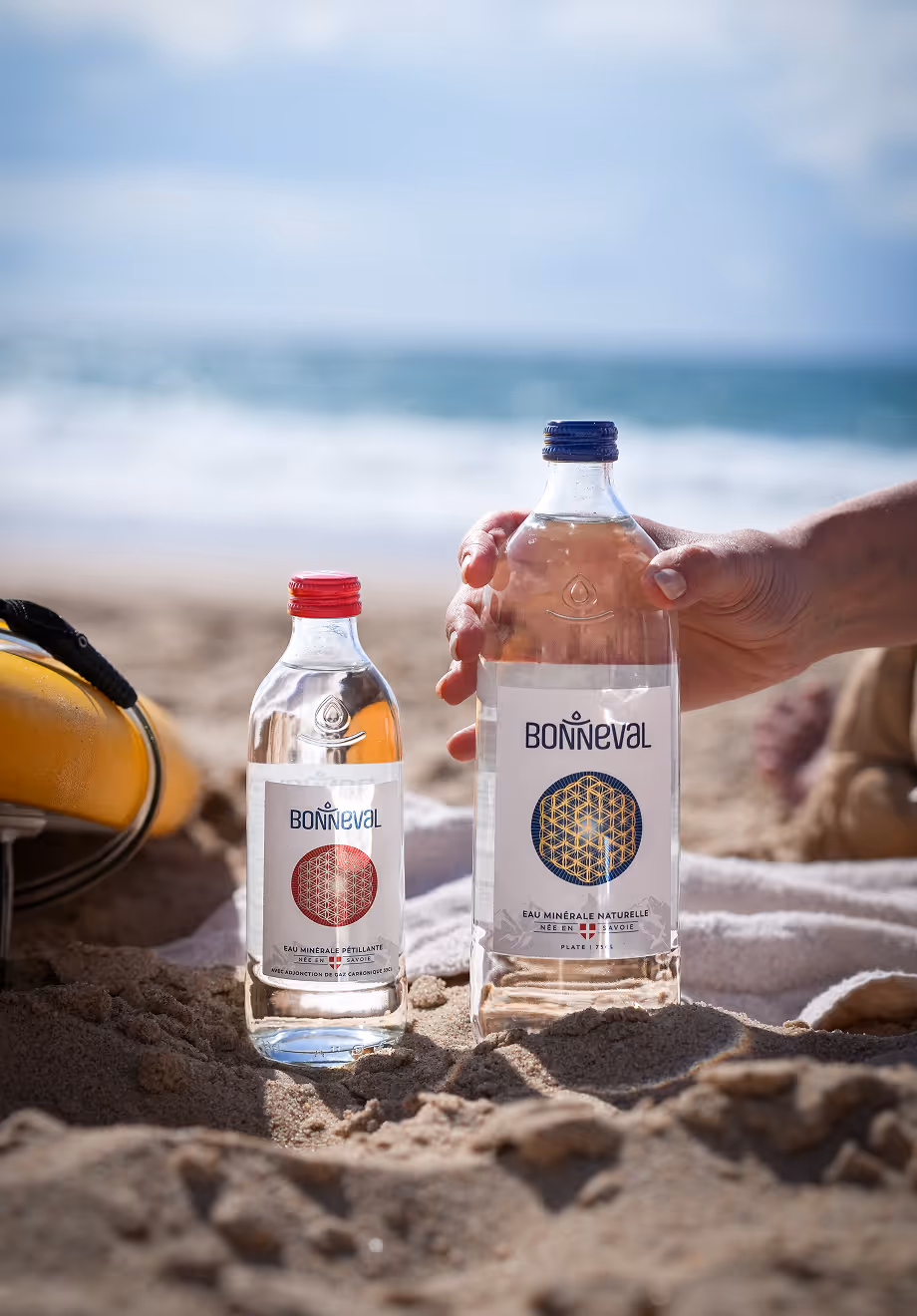 Deux bouteilles d'eau Bonneval posées sur le sable en bord de mer, une main tient la bouteille plus grande.