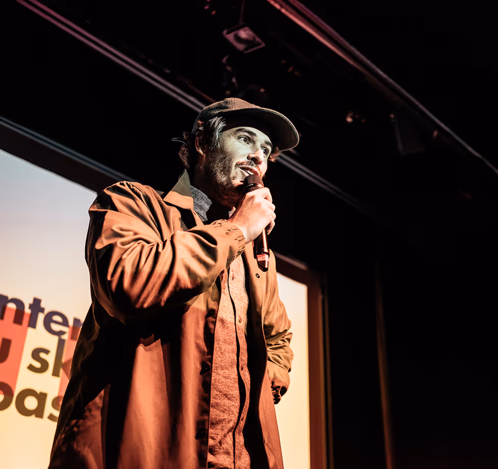 Un homme avec une casquette parle dans un micro sur scène devant un écran lumineux.