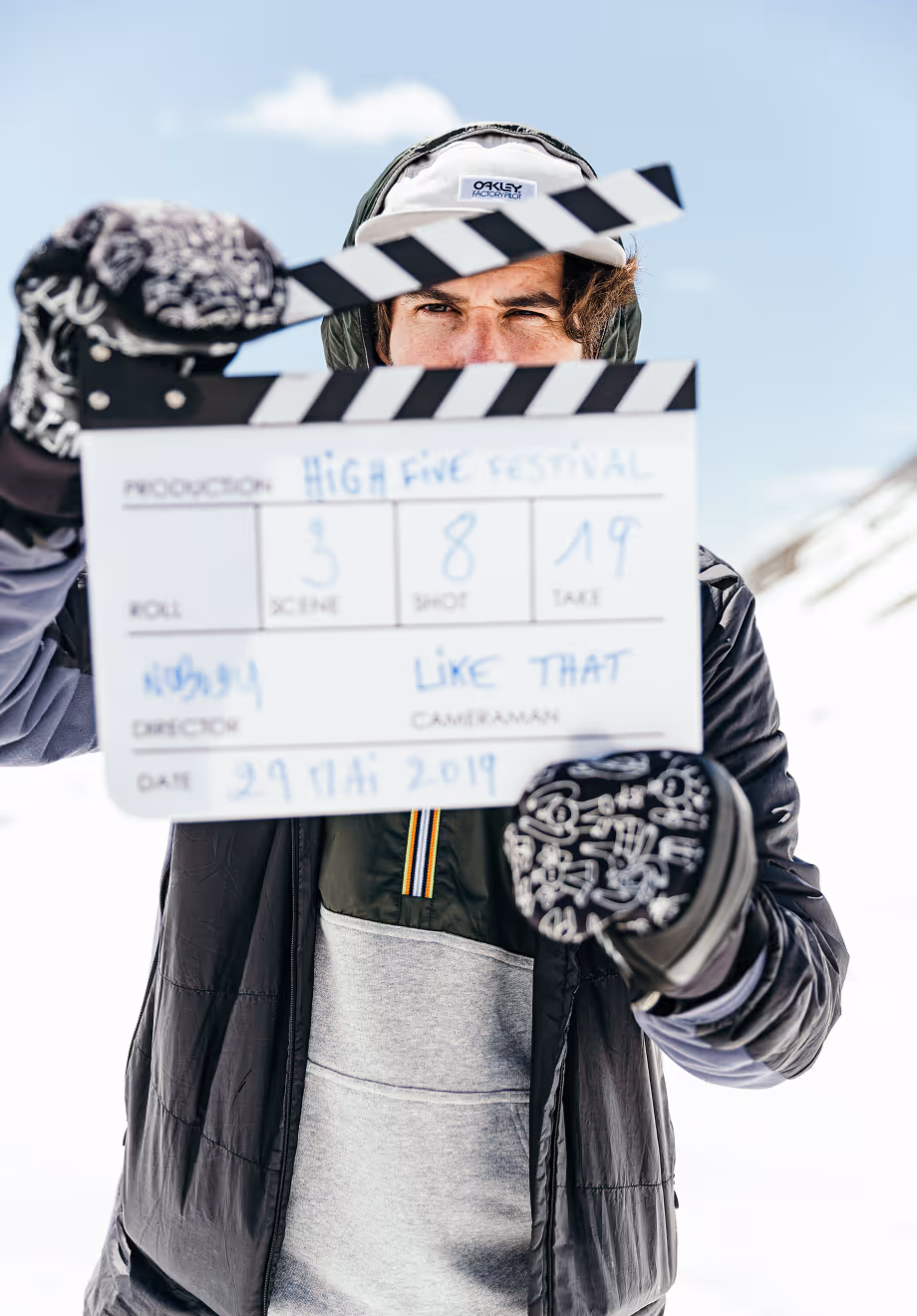 Une personne habillée en vêtements d'hiver tient une clap de cinéma marquée « High Five Festival » en montagne enneigée.