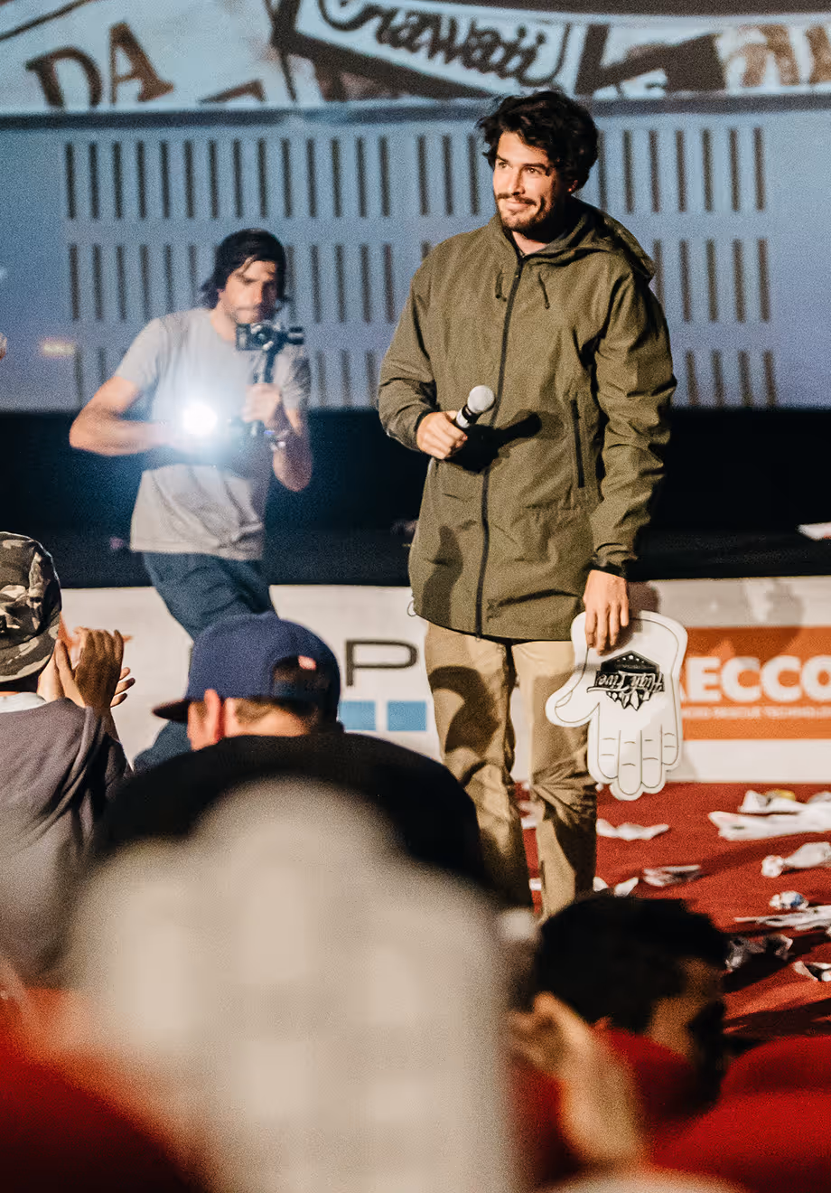 Un homme debout sur scène tenant un micro et un gant en mousse géant, avec un cameraman en arrière-plan et un public au premier plan.