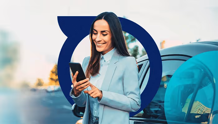 Smiling woman in a light blue blazer standing near a car, using a smartphone.