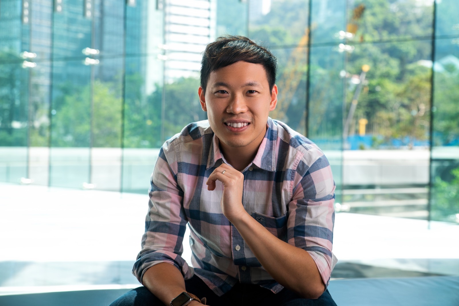 Smiling young man wearing a plaid shirt sitting indoors with a glass window and green trees in the background.