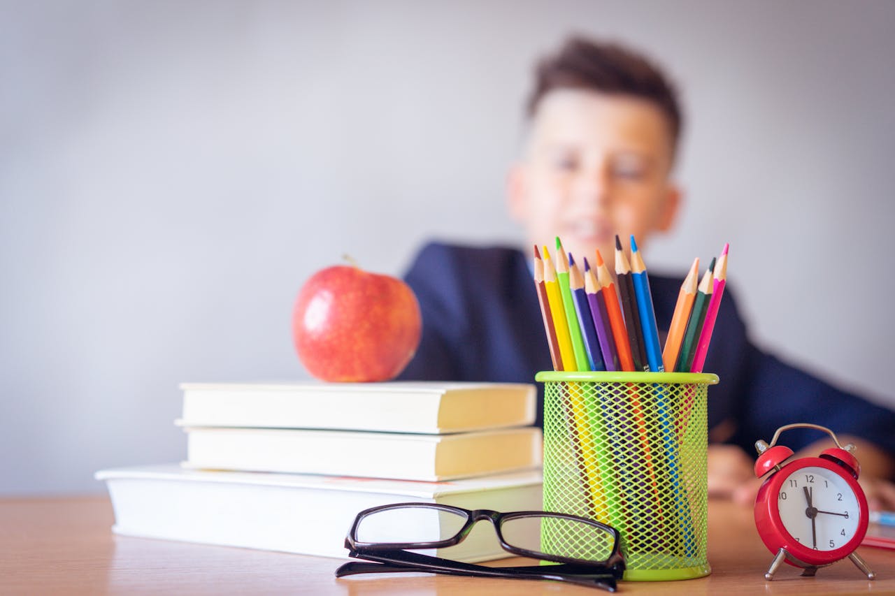 Stack of books with an apple on top, colorful pencils in a green holder, glasses, and a red alarm clock on a table with a blurred child in the background.