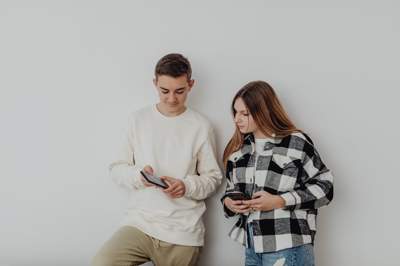 Teen boy and girl standing against a wall, looking at their smartphones.