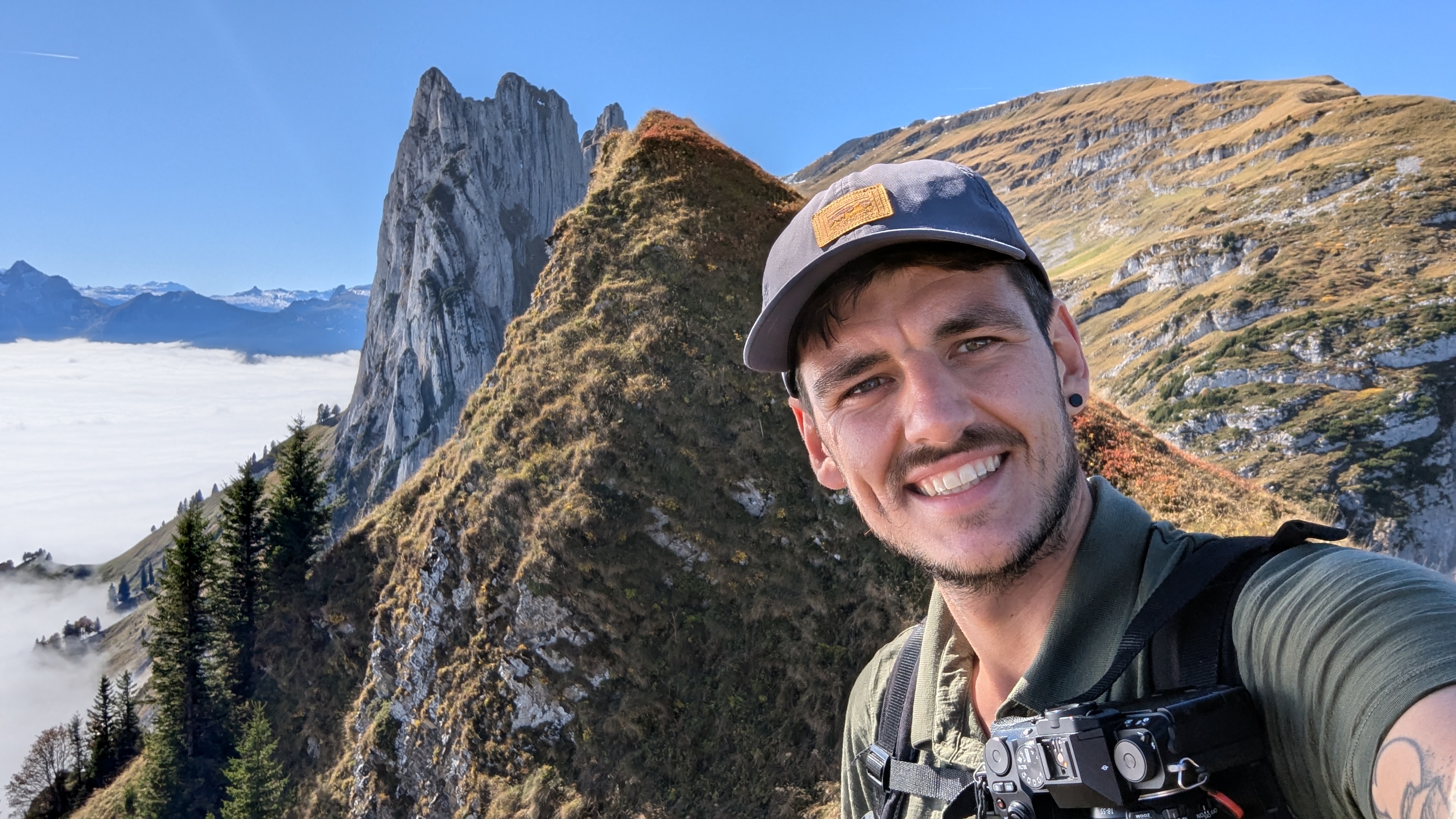 Mann mit Basecap lächelt in die Kamera vor einer felsigen Berglandschaft mit Nadelbäumen und Nebel darunter.