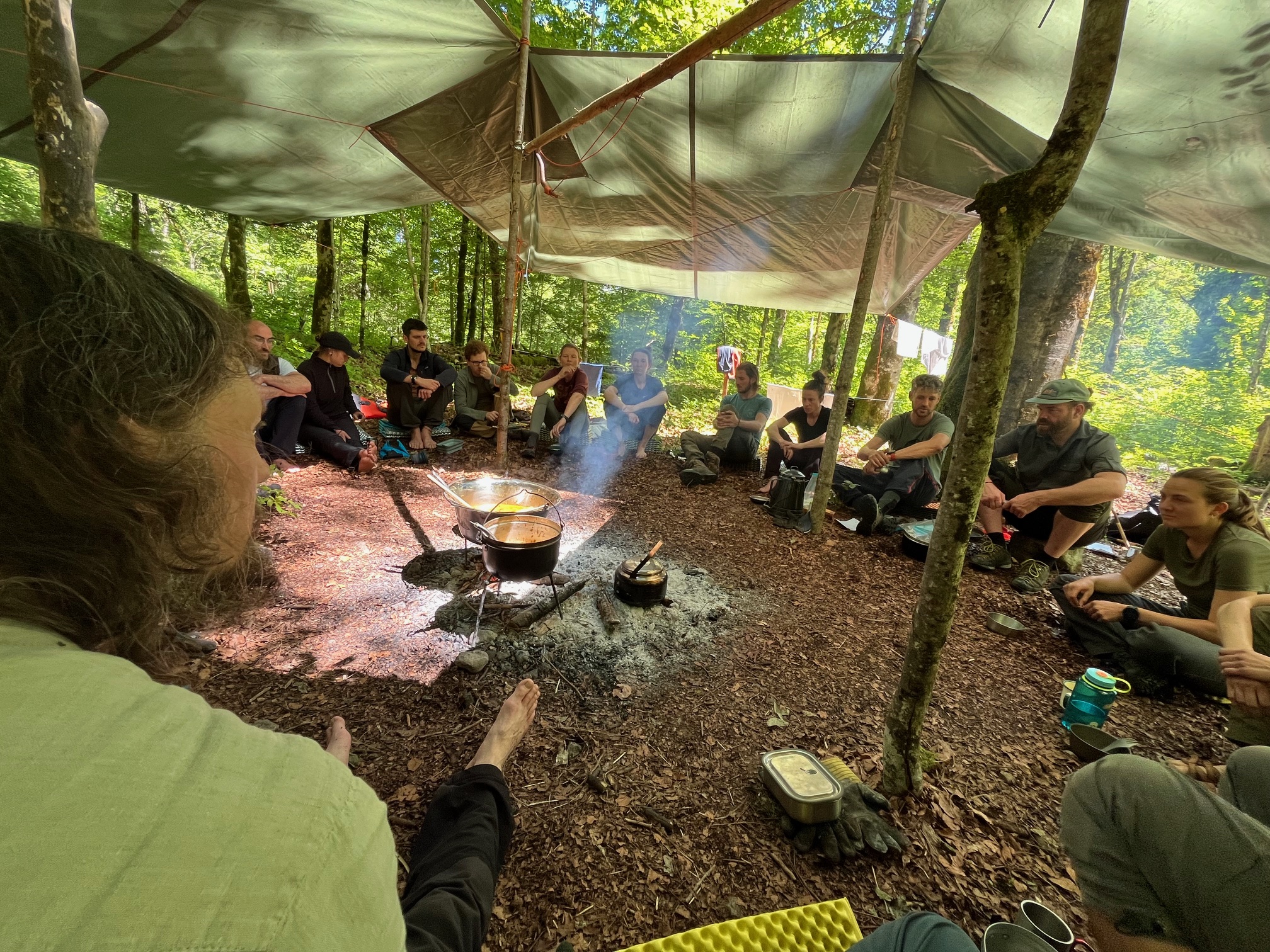 Menschen sitzen im Wald auf dem Boden unter einem Zeltdach rund um ein Lagerfeuer mit Kochtöpfen.