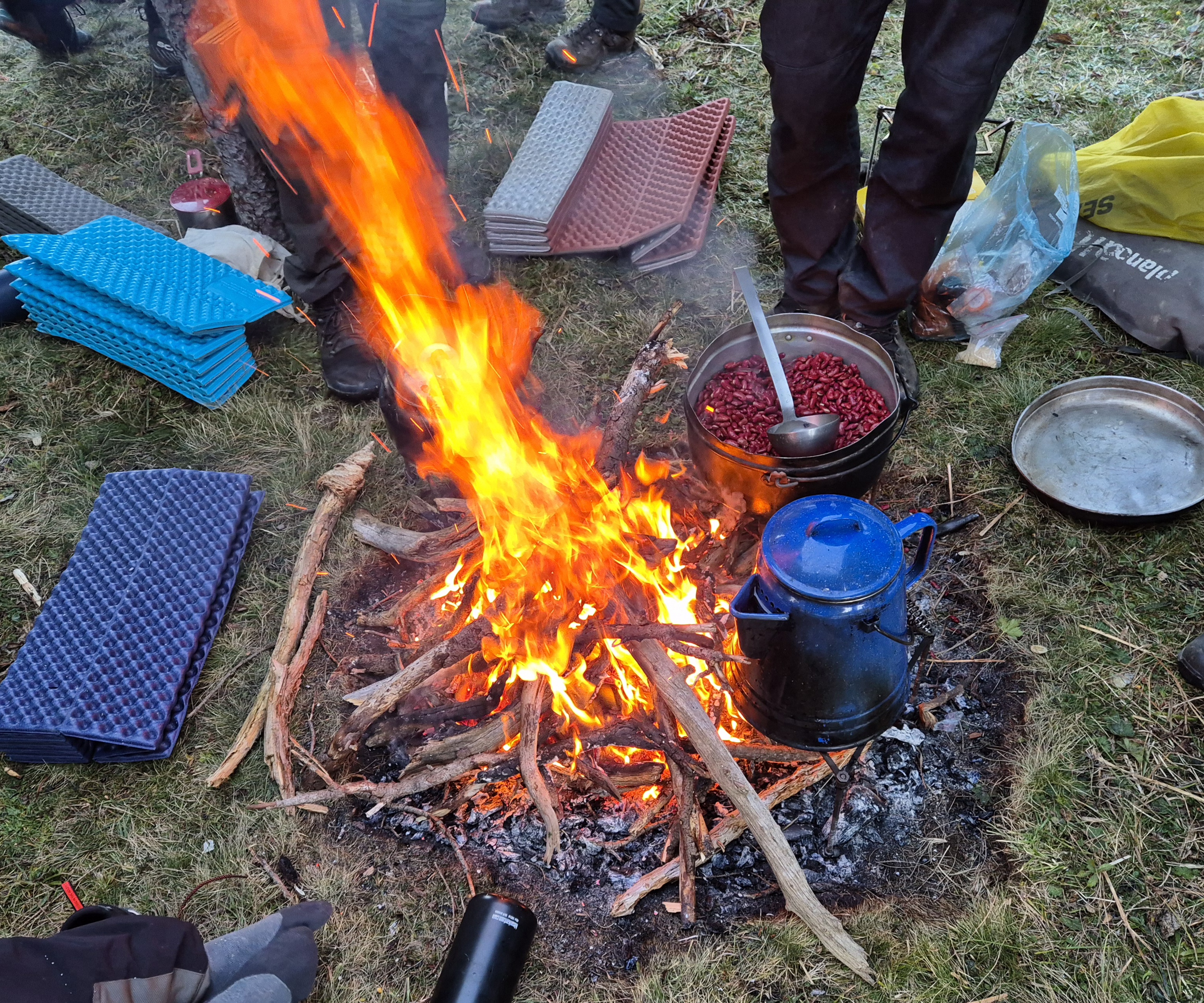 Lagerfeuer mit Kochtopf voller roten Bohnen und blauer Kanne, Personen stehen darum, Camping-Schlafmatten liegen auf Gras.