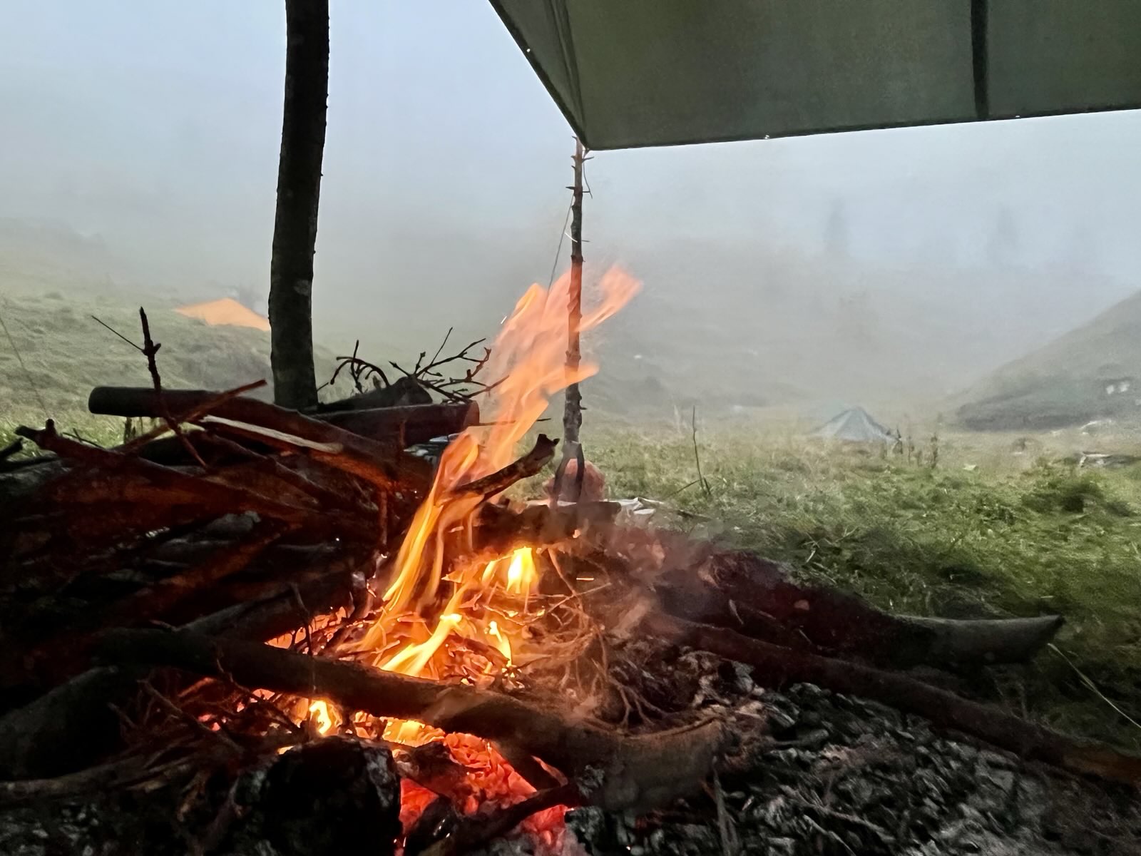 Feuer mit brennendem Holz unter einem Zeltdach in einer nebligen, grasbewachsenen Landschaft.