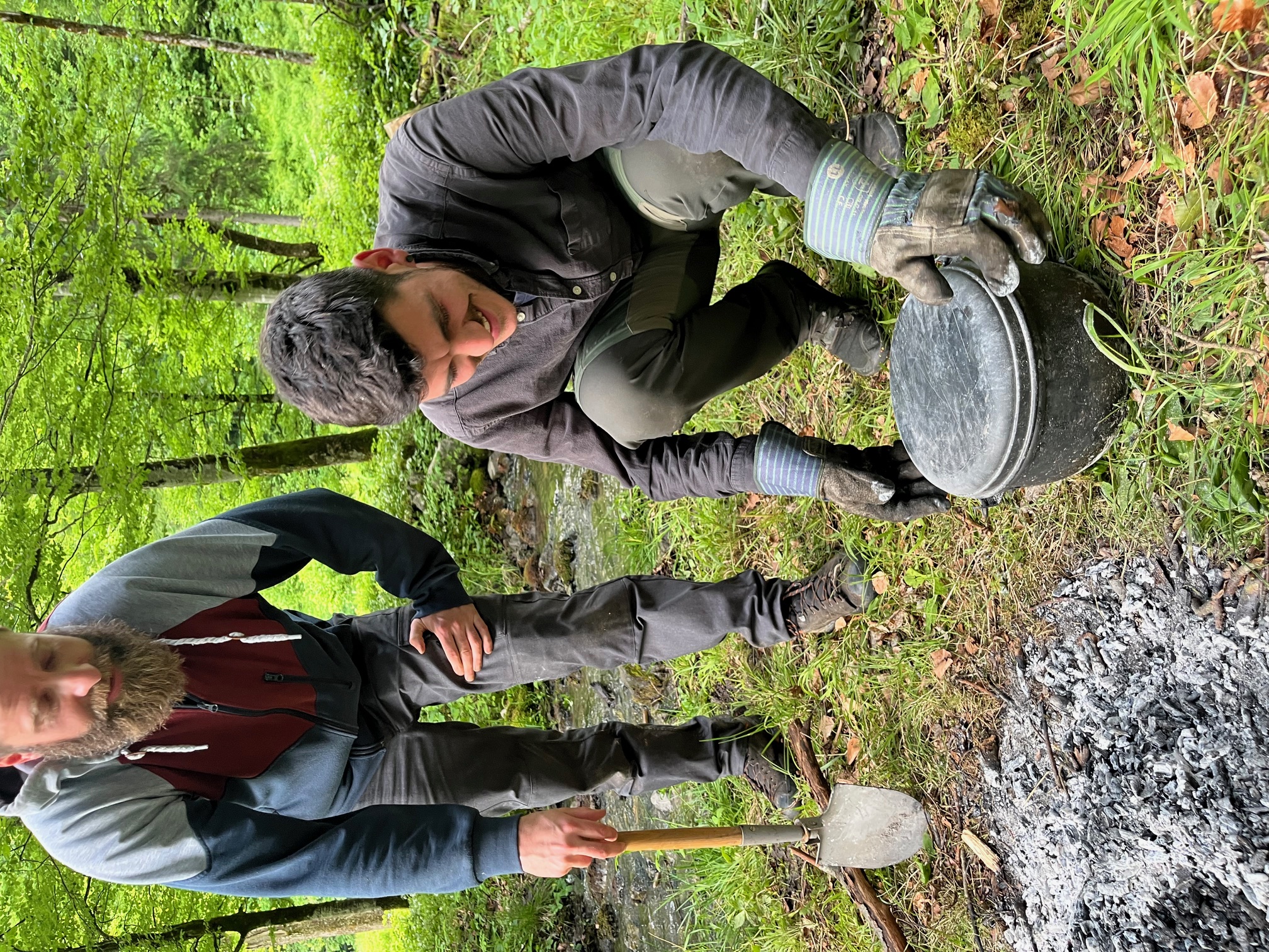 Zwei Männer im Wald beim Kochen über einem Lagerfeuer, einer hockt und hält einen Topf mit Ofenhandschuhen, der andere steht mit einer kleinen Schaufel.