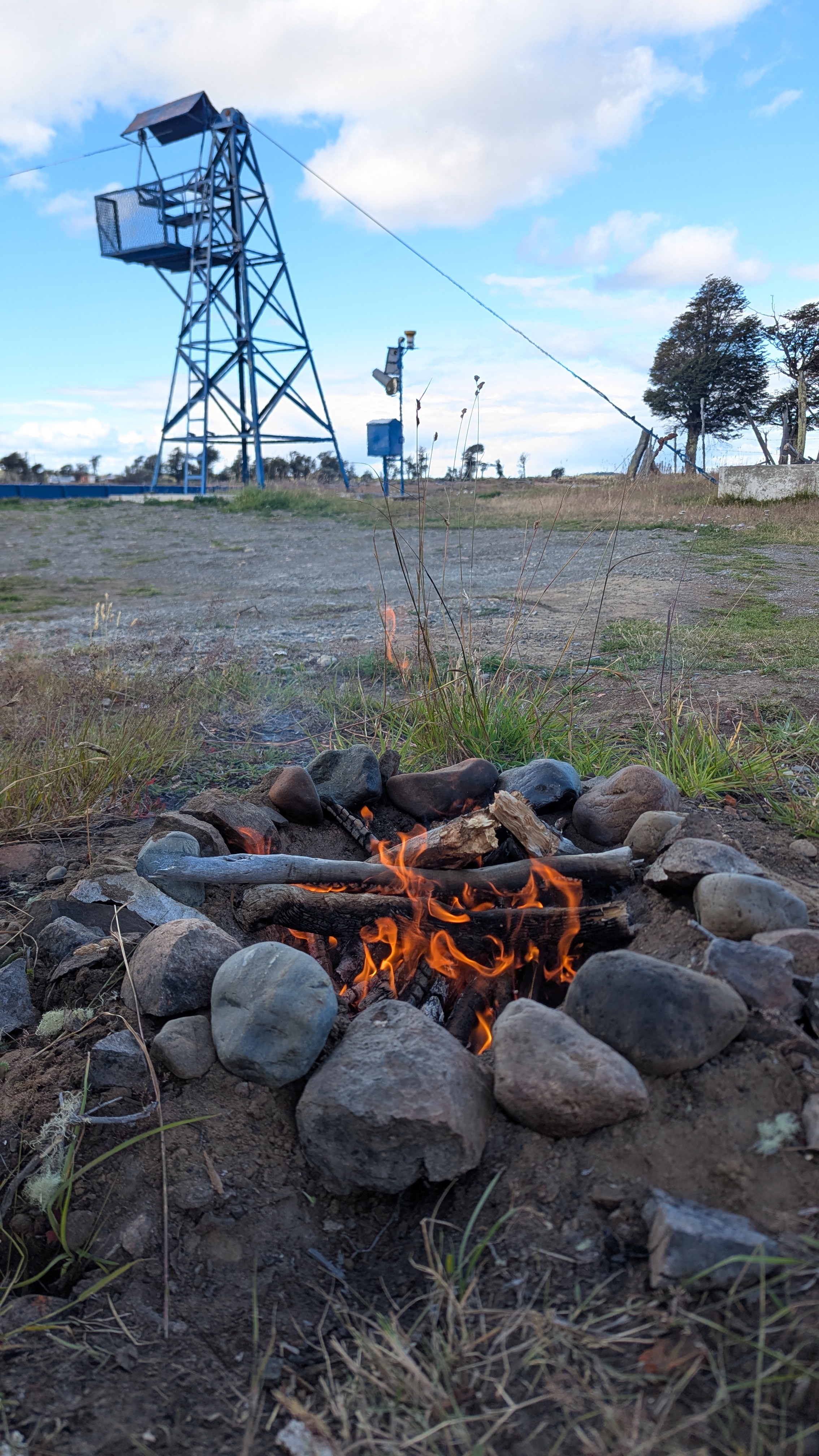 Lagerfeuer umgeben von Steinen auf offenem Gelände mit einem Aussichtsturm und Blauem Himmel im Hintergrund.