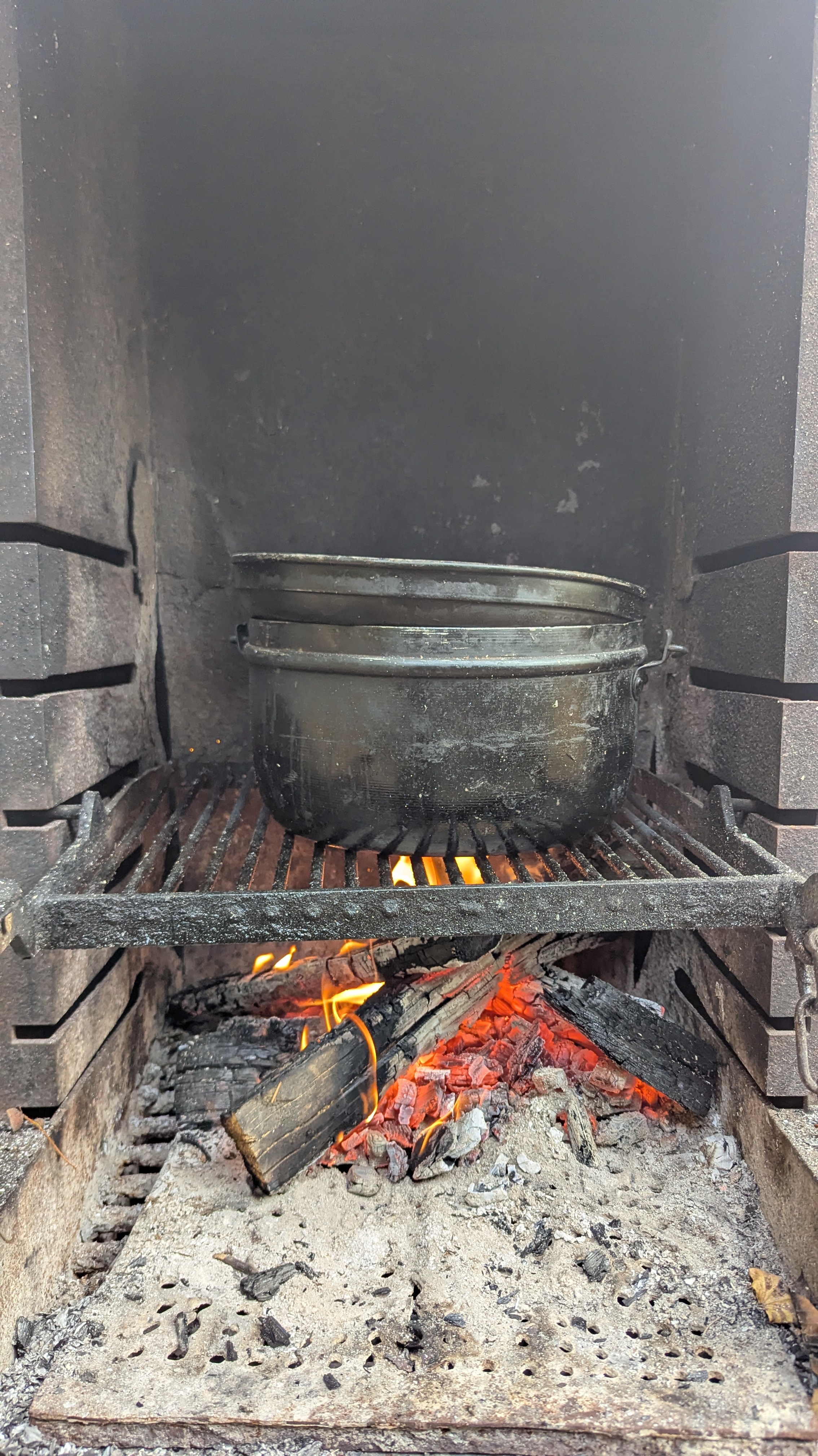 Schwarzer Topf auf einem schwarzen Grillrost über glühenden Holzscheiten mit Flammen in einem gemauerten Ofen.