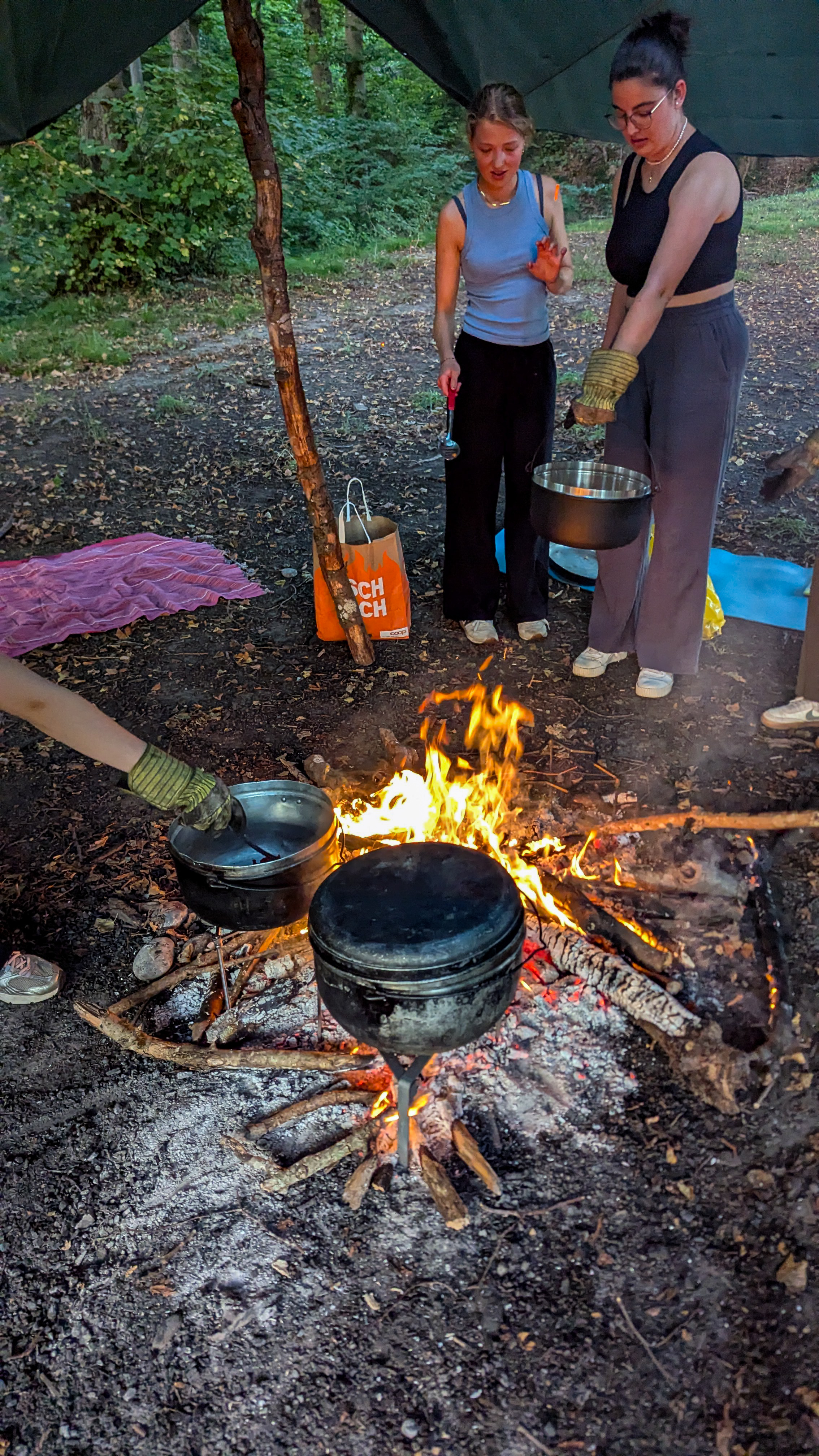Zwei Frauen kochen über offenem Feuer im Wald, eine hält einen Topf mit Ofenhandschuhen, während ein anderer Topf auf dem Feuer steht.