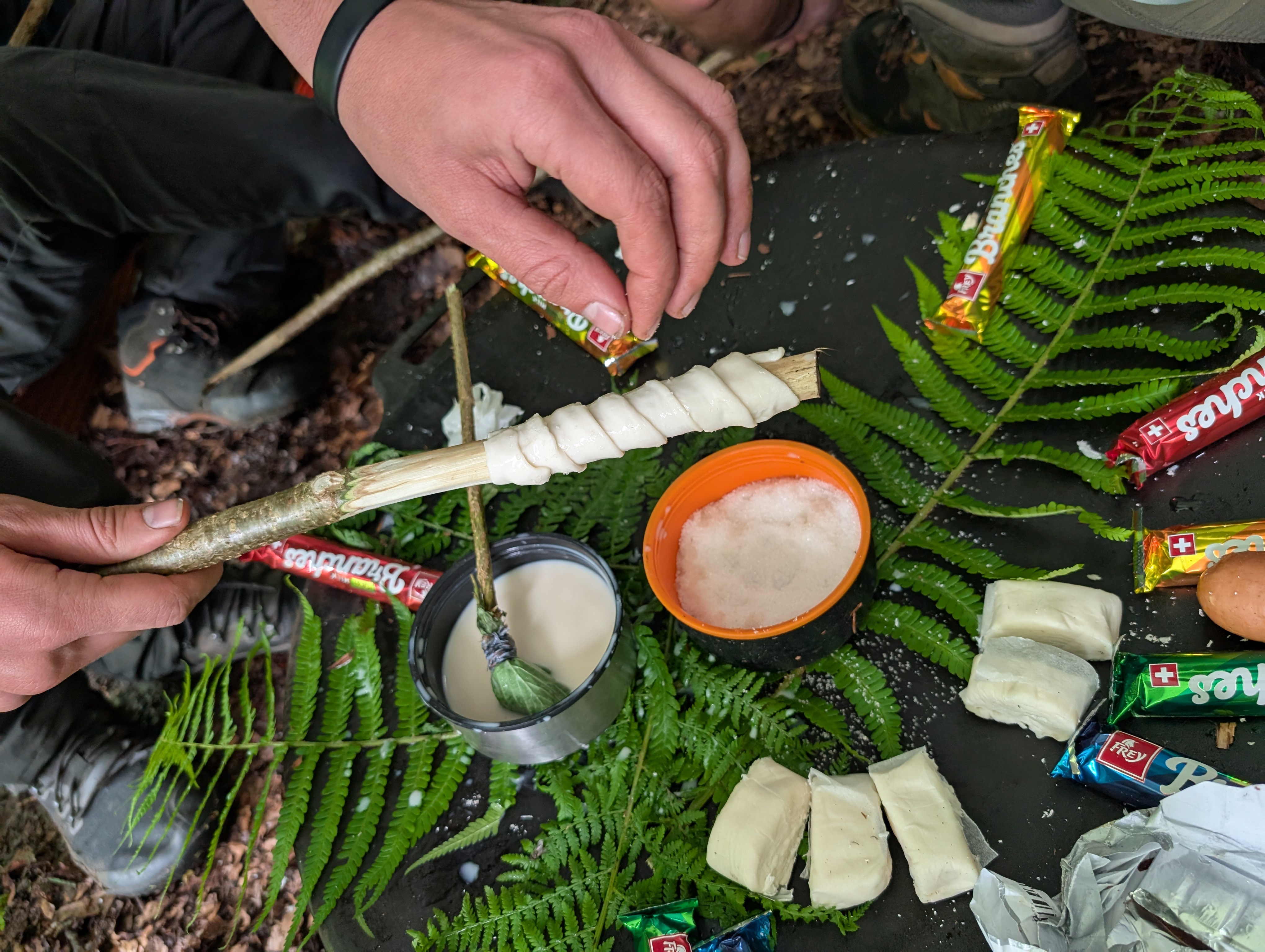 Person wickelt Teig um einen Stock, um ihn über Feuer zu backen, umgeben von Brenntal-Schokoladen, einem Ei, Schalen mit Flüssigkeiten und Farnblättern.