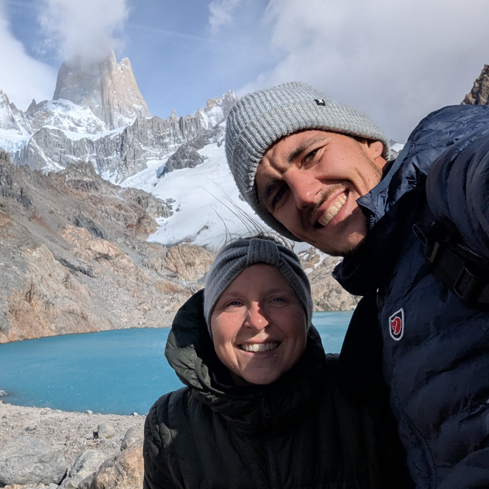 Zwei Personen in Winterbekleidung vor einem Bergsee und schneebedeckten Gipfeln.