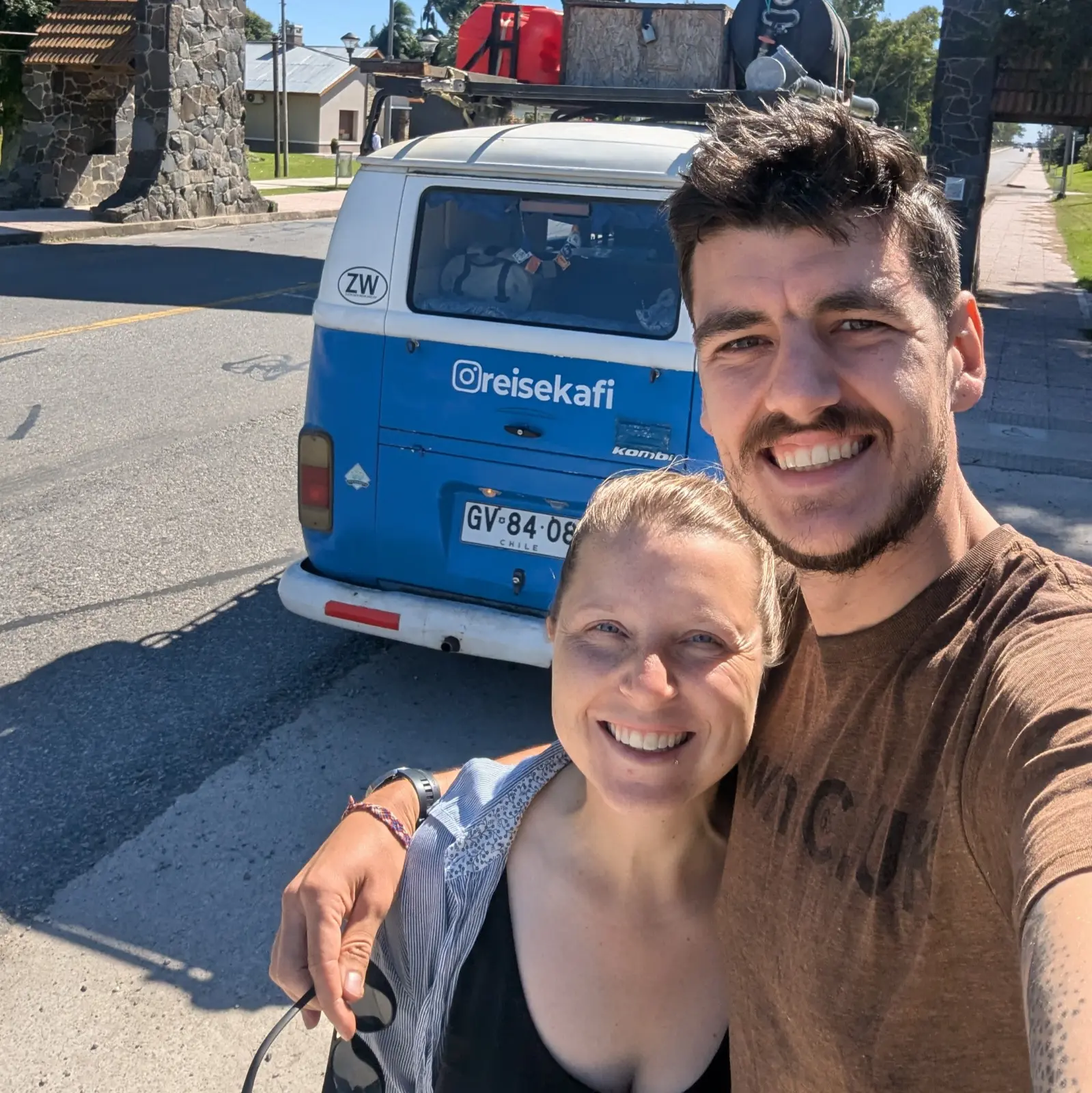 Ein lächelndes Paar macht ein Selfie vor einem blauen und weißen VW-Bus mit Instagram-Namen @reisekafi auf der Rückseite.