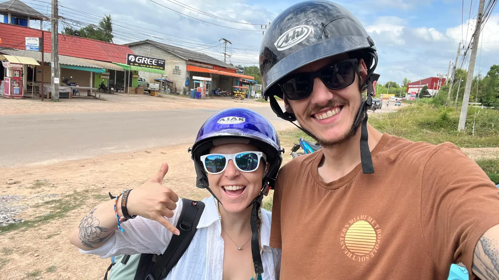 Fröhliches Paar mit Helmen und Sonnenbrillen macht Selfie auf einer Straße mit Geschäften im Hintergrund.