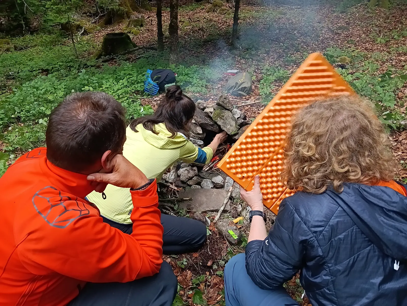 Drei Personen, die ein Lagerfeuer im Wald vorbereiten, eine davon hält eine orange Isomatte.
