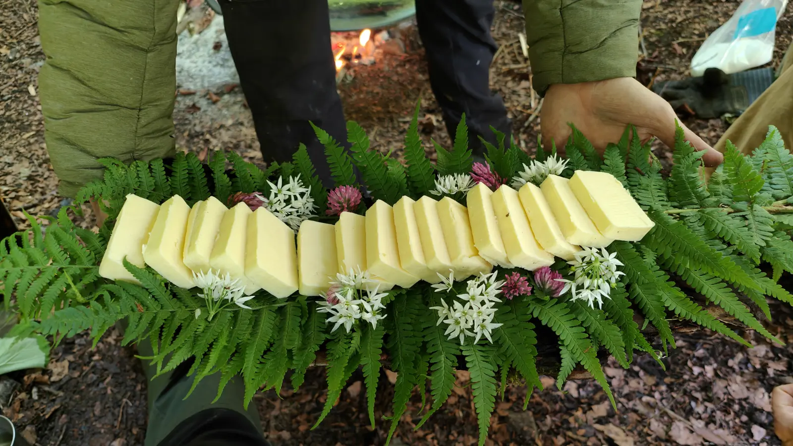 Vorgeschnittene Butterstücke auf Farnblättern mit weißen und rosa Wildblumen, gehalten von zwei Personen in einer Waldumgebung.