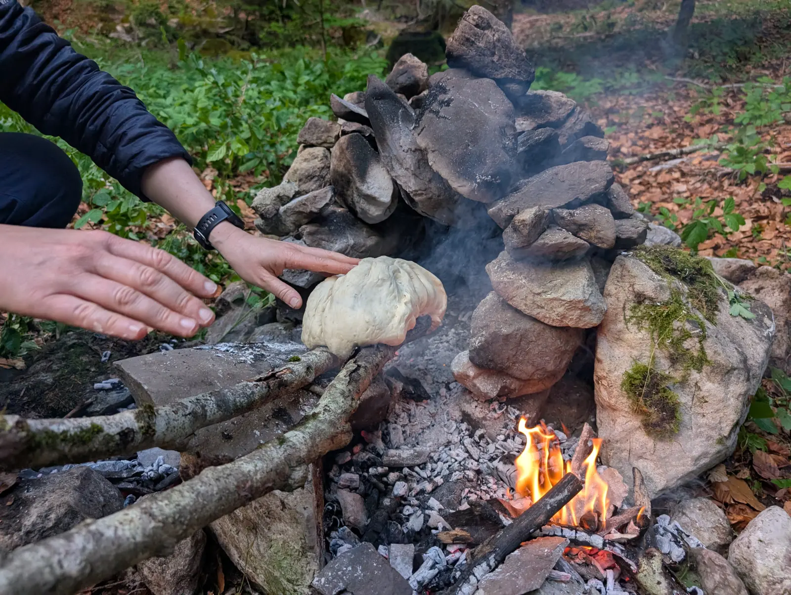 Person bäckt Brot über einem offenen Feuer in einer Steinfeuerstelle im Wald.