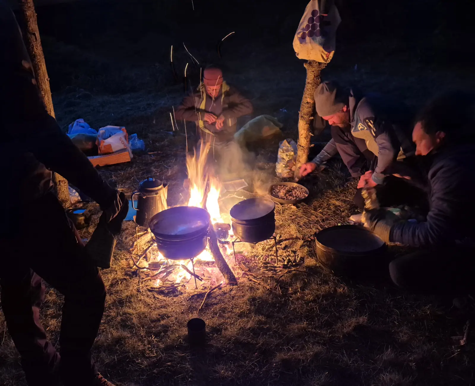 Drei Personen kochen in der Nacht auf offenem Feuer im Freien, mit Töpfen und Pfannen über der Glut.
