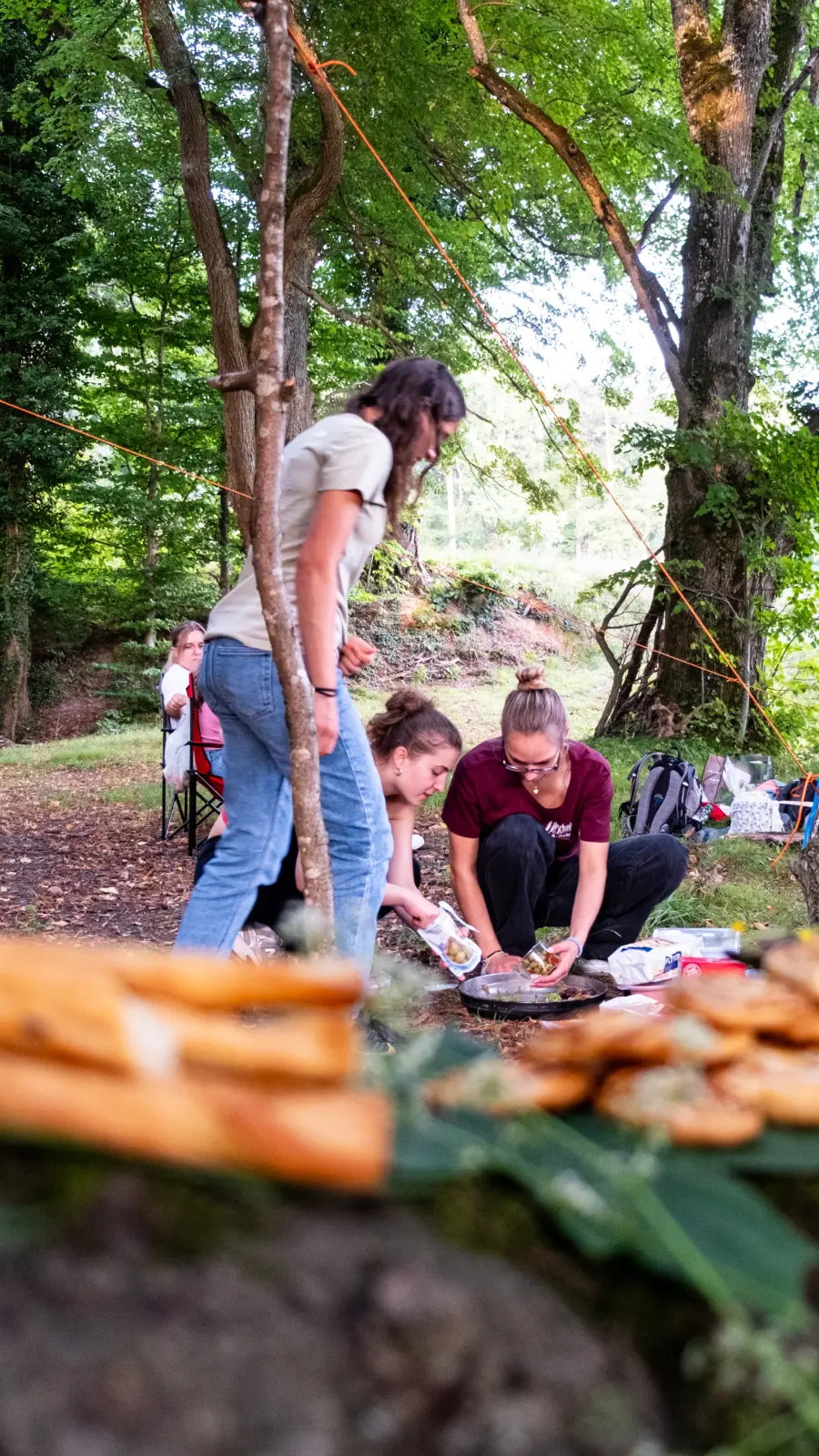Drei Frauen bereiten im Wald eine Mahlzeit auf dem Boden zu, während eine vierte Frau auf einem Campingstuhl im Hintergrund sitzt.