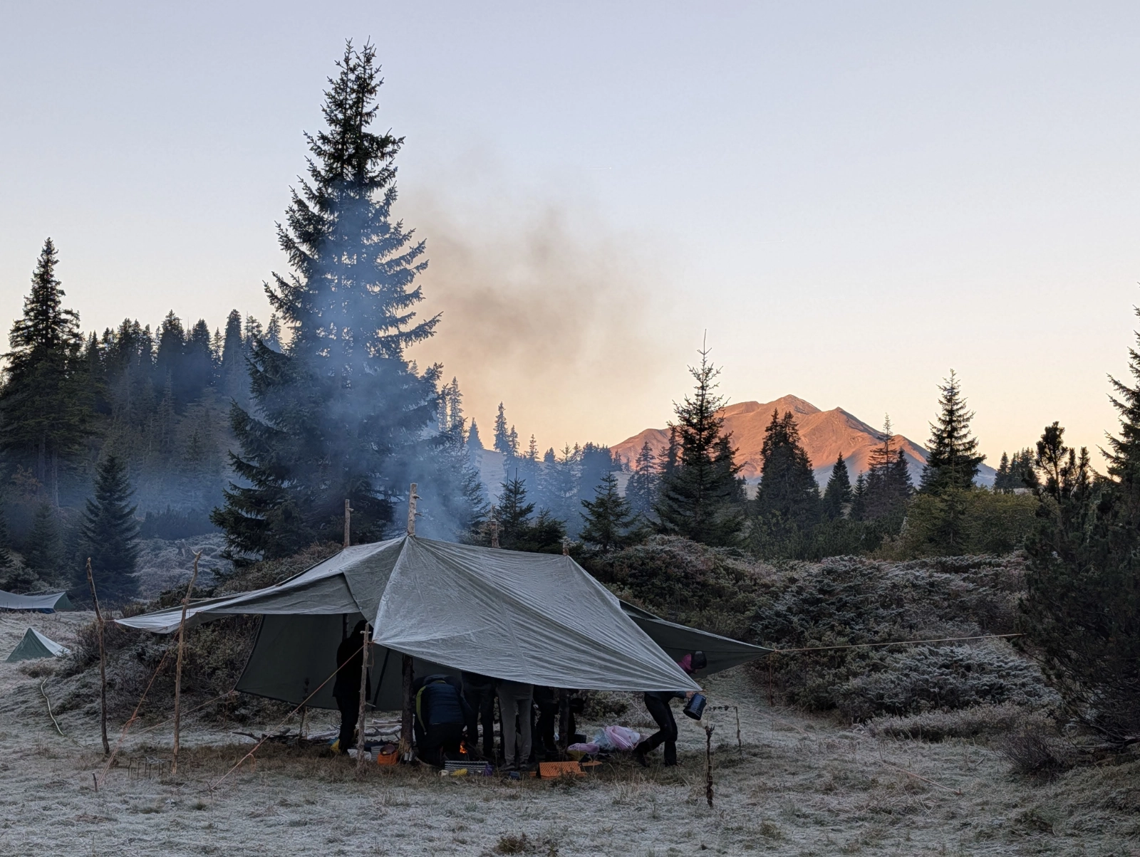 Zeltgruppe im Wald auf frostbedecktem Boden mit Rauch, der in den Himmel steigt, und Bergen im Hintergrund bei Sonnenaufgang.