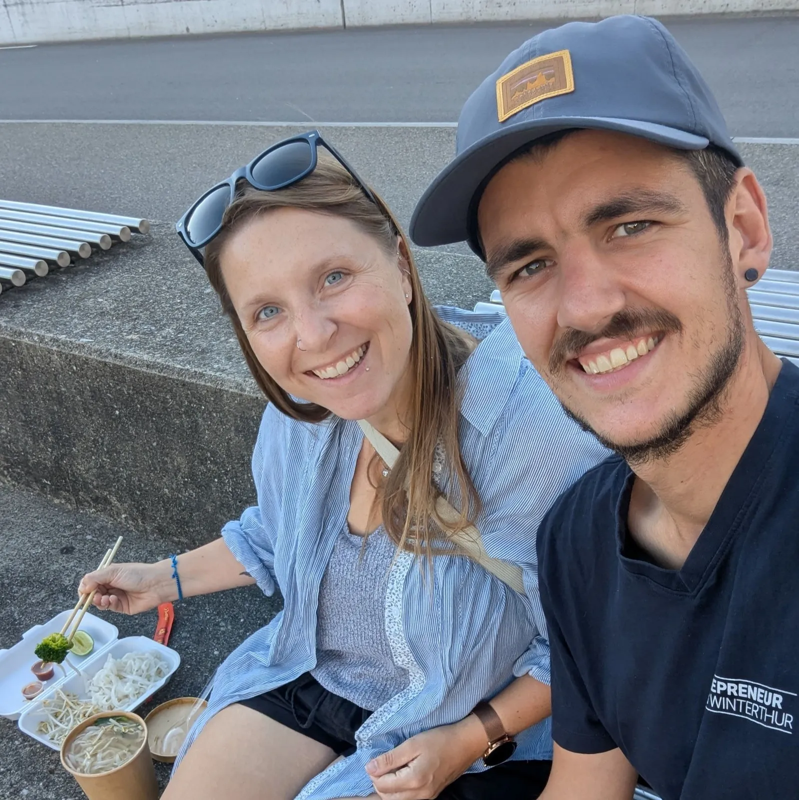 Ein lächelndes Paar sitzt auf einer Bank im Freien und isst asiatische Nudeln und Gemüse.