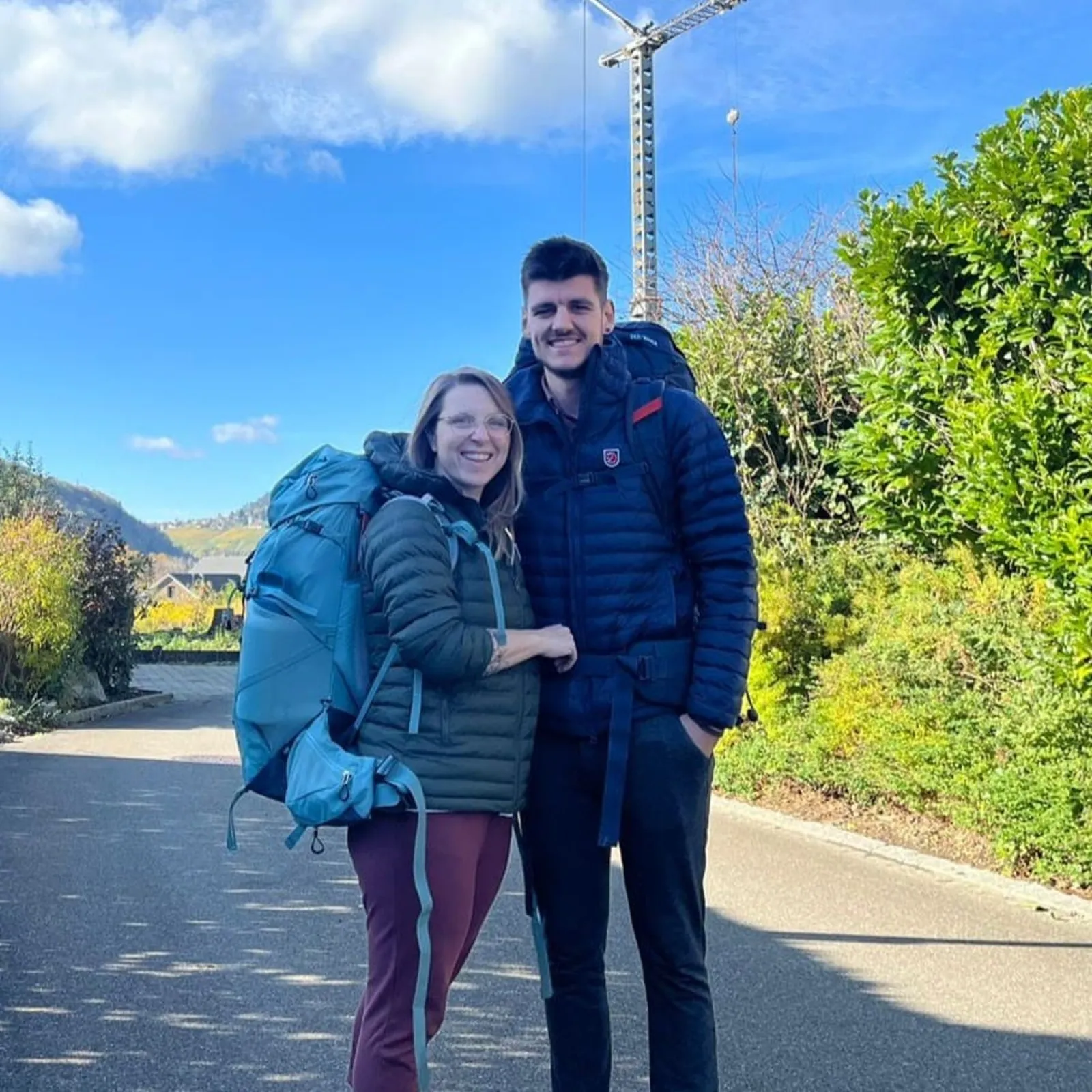 Zwei Wanderer mit Rucksäcken posieren und lächeln auf einem sonnigen Weg mit grünem Laub im Hintergrund.
