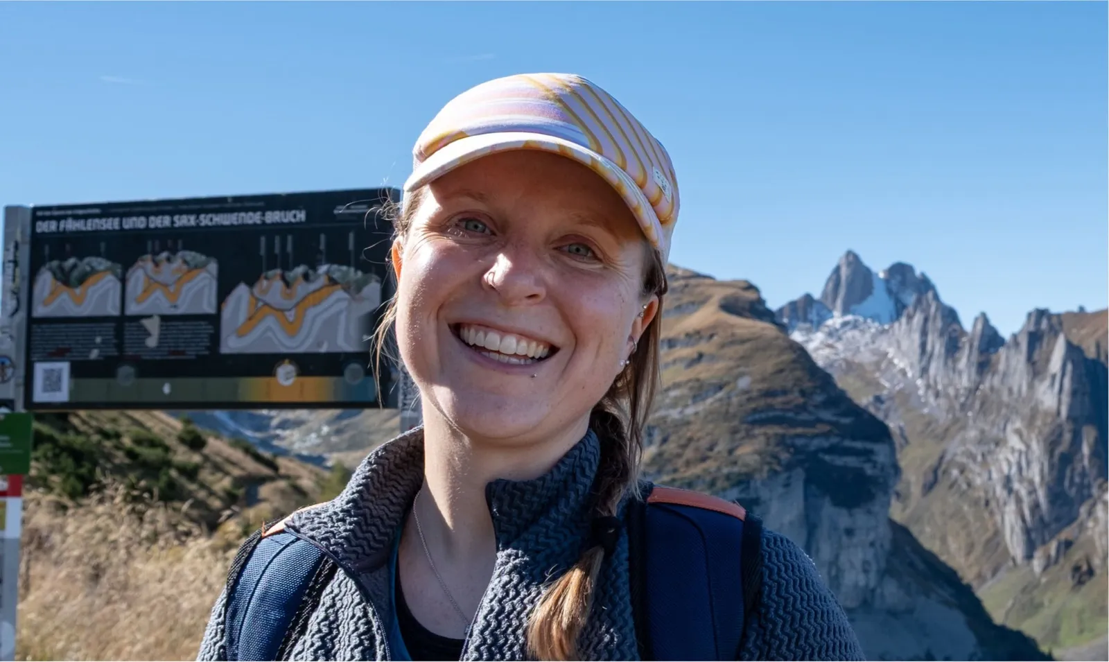 Lächelnde Frau mit Cap und Wanderkleidung vor Berglandschaft mit Informationstafel.