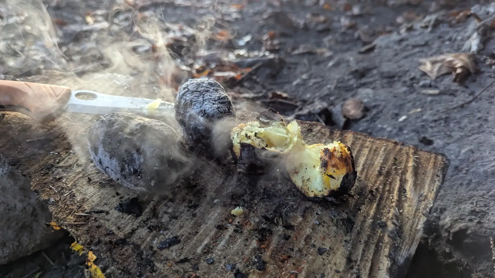 Heiße, angebrannte Kartoffeln mit Dampf auf einem Holzbrett im Freien, daneben ein Messer mit Holzgriff.