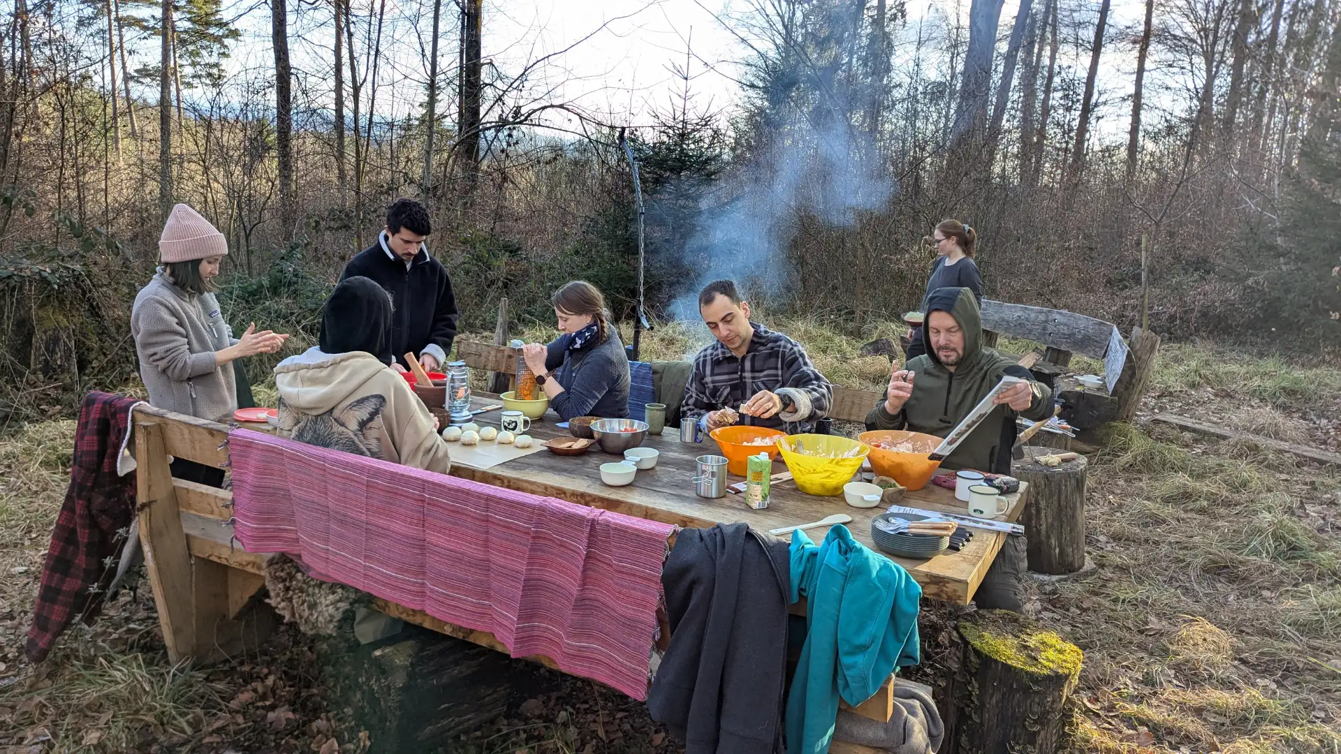 Sechs Personen sitzen im Freien an einem Holztisch und bereiten gemeinsam Essen zu, umgeben von Bäumen und Gras.
