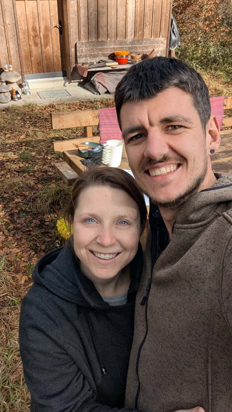 Ein lächelndes Paar umarmt sich vor einem Holztisch und einer Holzscheune im herbstlichen Garten.