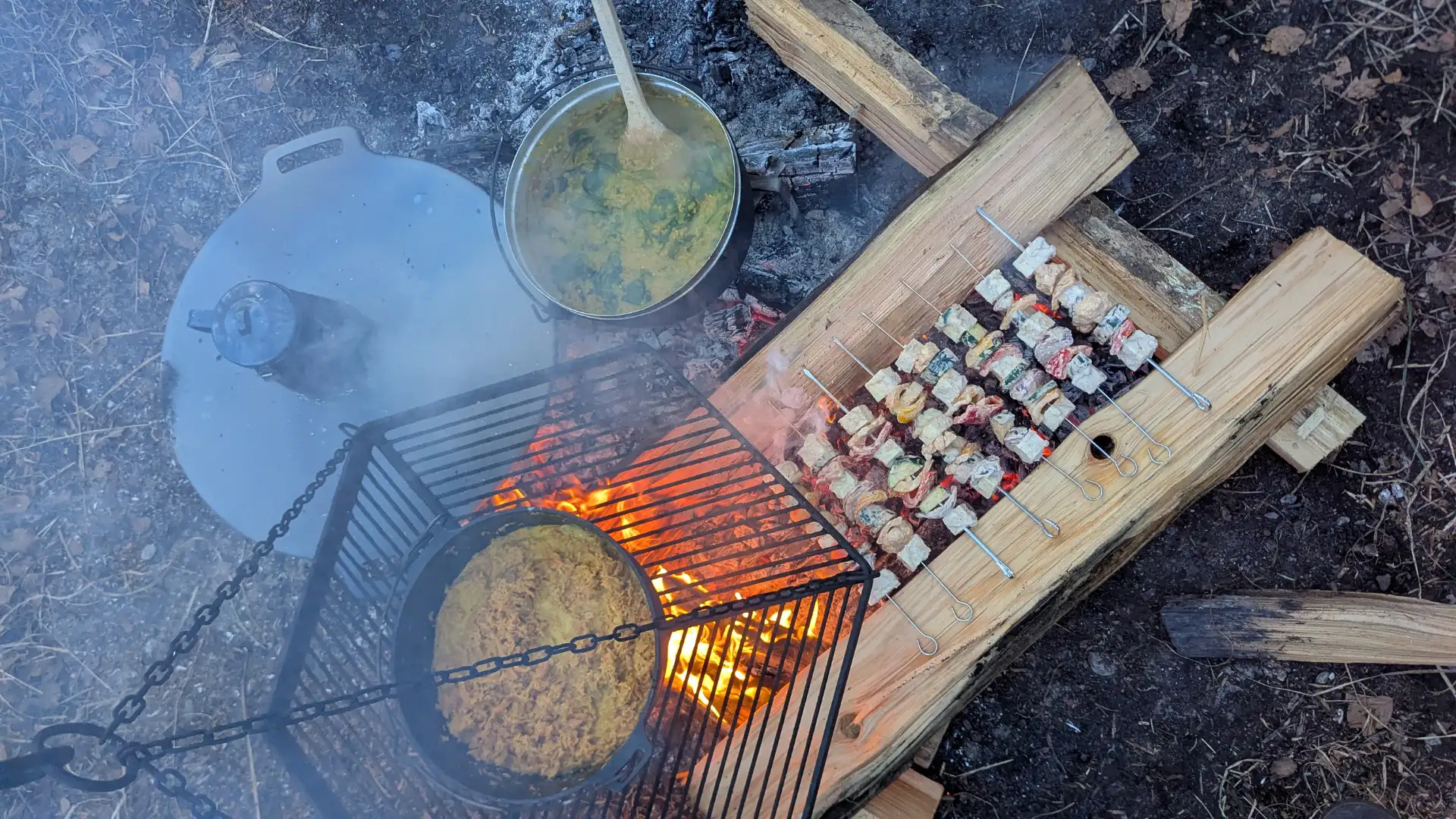 Feuerstelle mit Grillrost und Topf, dazu Gemüse-Spiesse auf Holzstücken über glühender Glut.