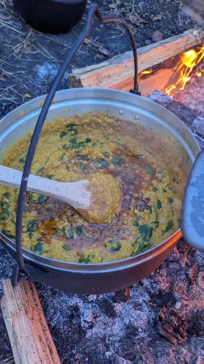 Grosser Kochtopf mit gelbem Linsengericht und grünem Gemüse, der auf einem offenen Lagerfeuer mit Holzscheiten steht.