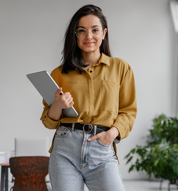 Young woman with glasses wearing a mustard yellow shirt and light jeans holding a tablet in an office setting.