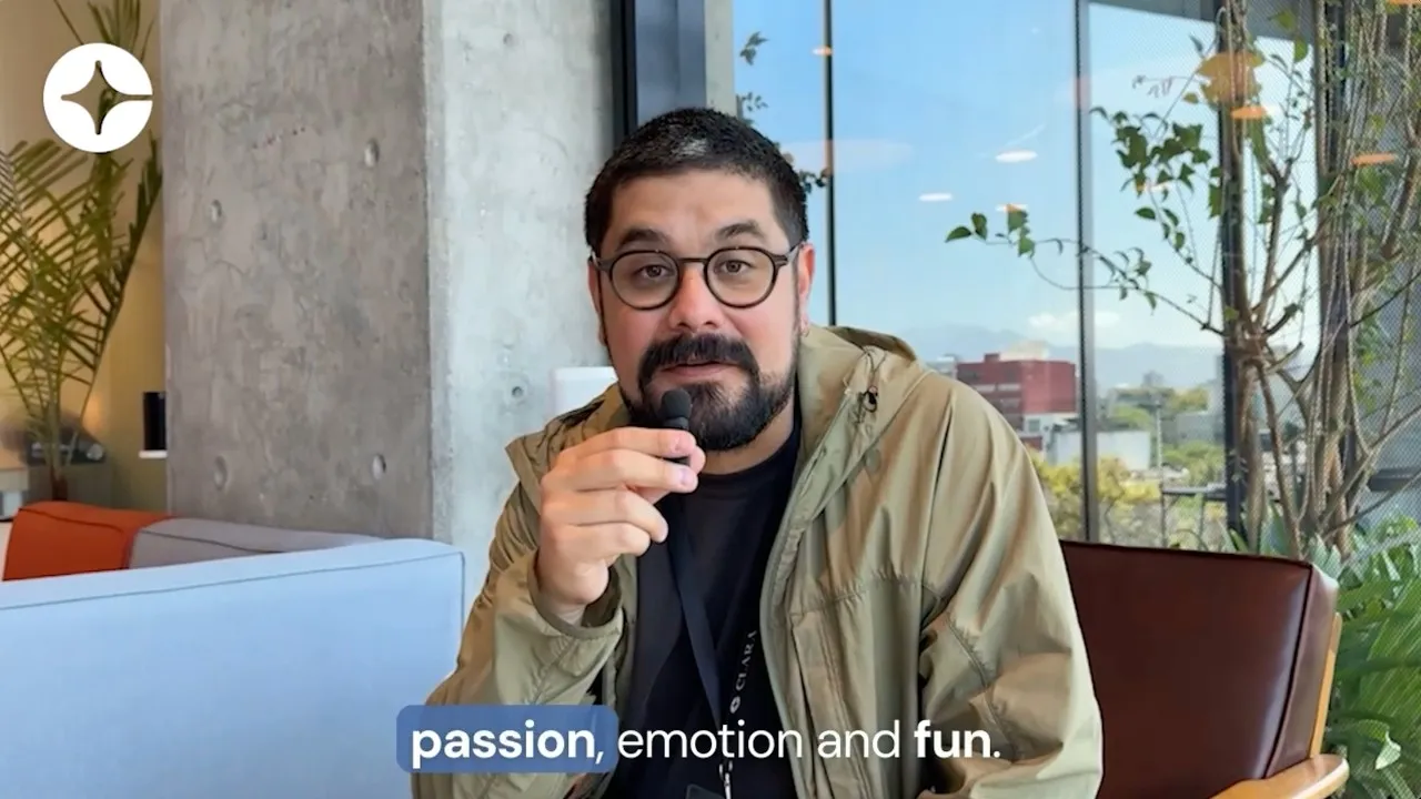 Man with beard and glasses speaking into a microphone in a modern office with plants and large windows.