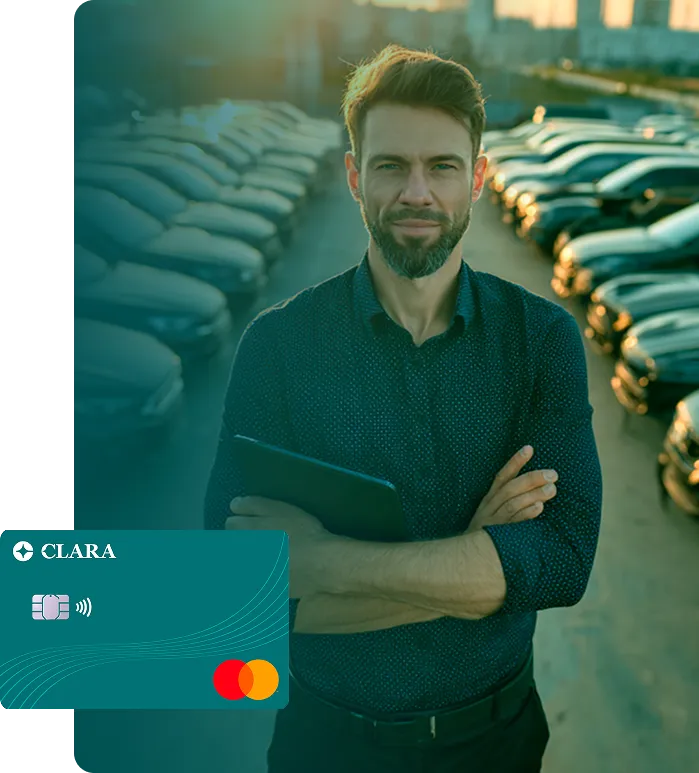 Man with beard holding tablet standing in car lot with rows of parked cars at sunset.