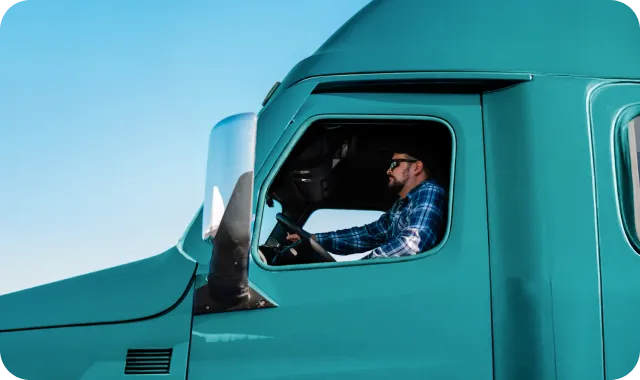 Man in sunglasses driving a teal semi-truck cab against clear blue sky.