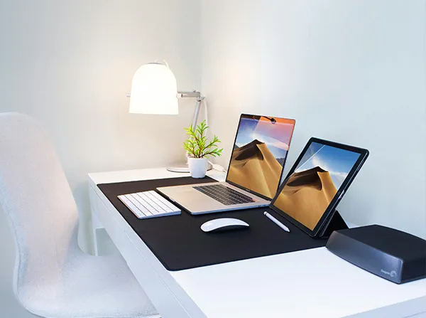 Minimal white desk setup with laptop, tablet, keyboard, mouse, lamp, plant, and a gray chair.