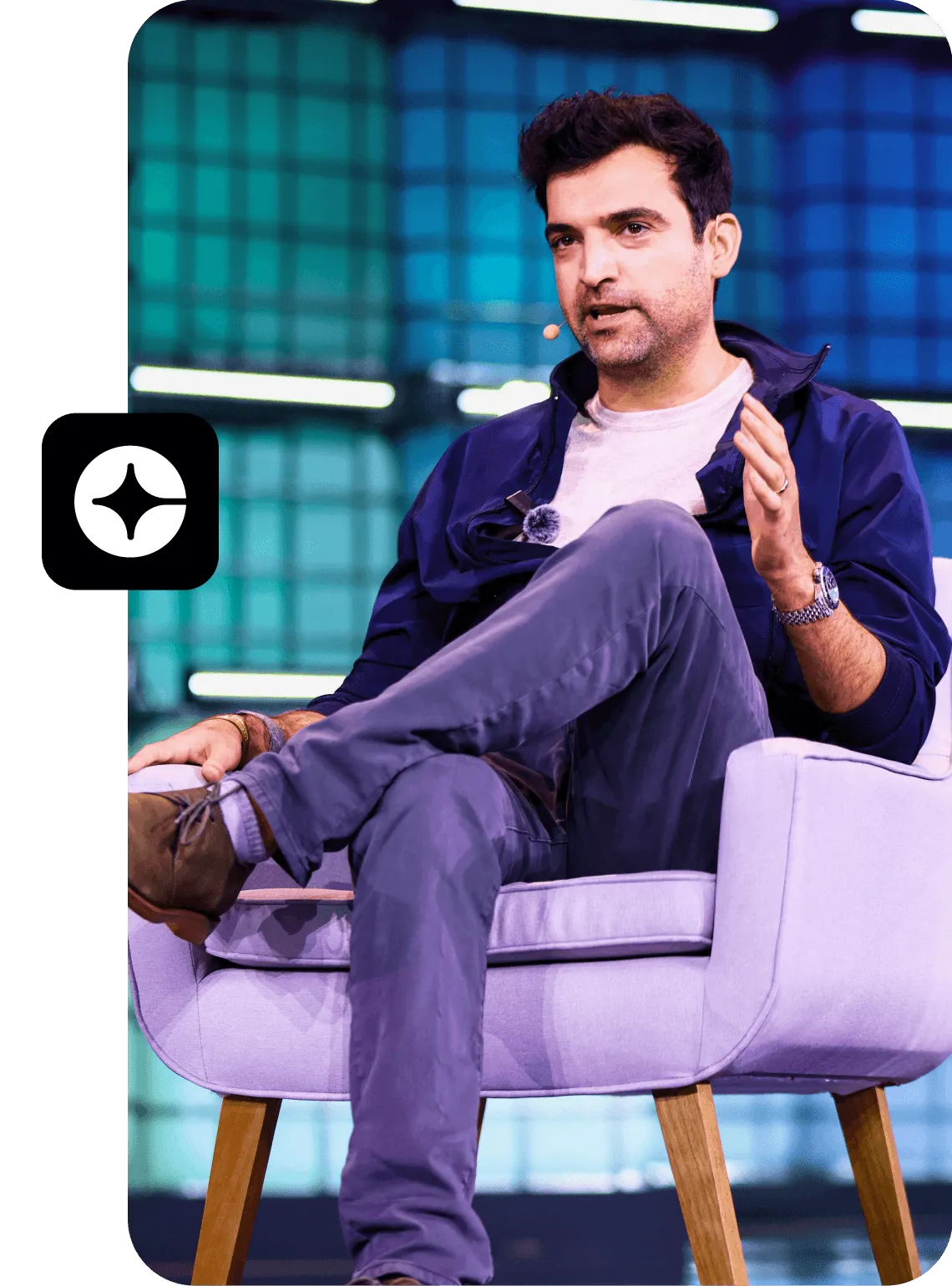 Man with microphone talking while seated on a purple chair with crossed legs at an event.