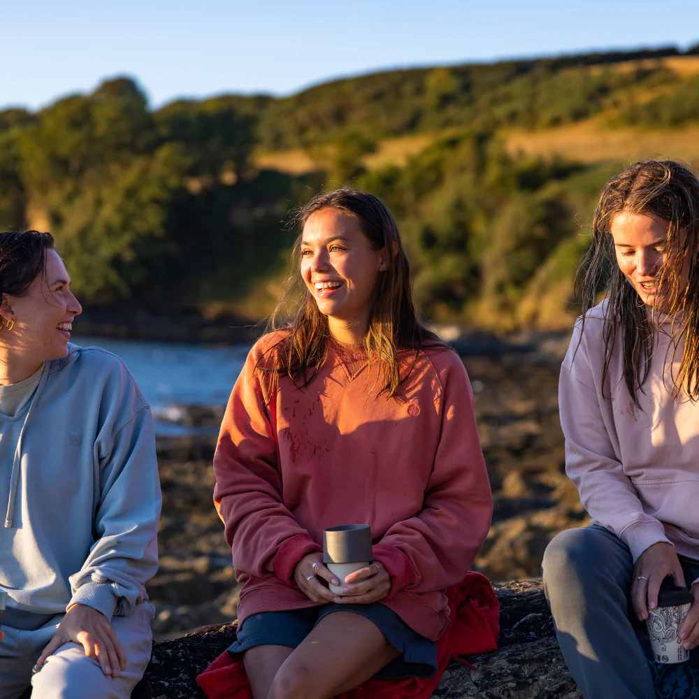 Three women sitting on rocks by the water, smiling and holding cups, with trees and hills in the background.