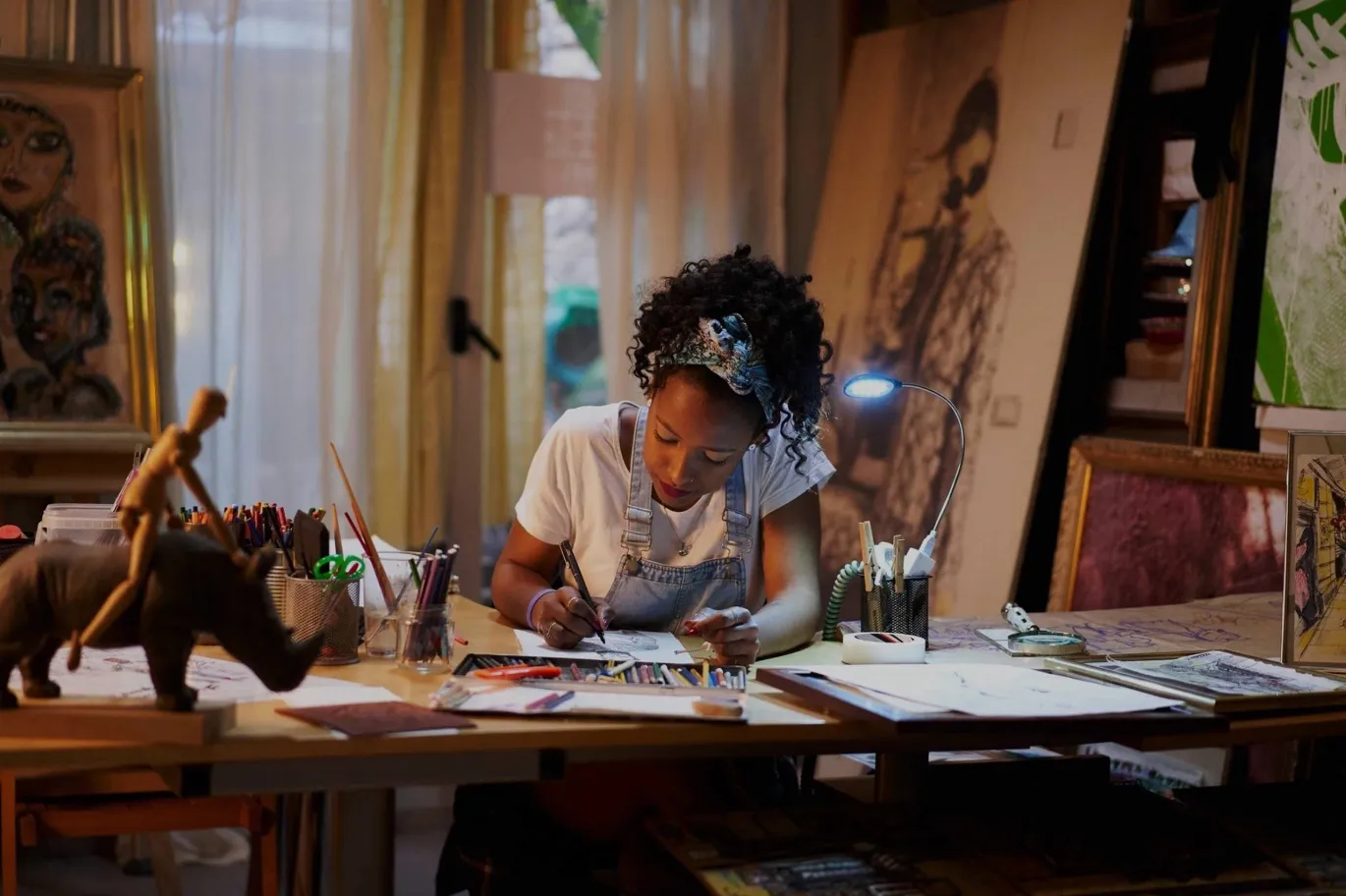 Woman with curly hair drawing at a desk surrounded by art supplies and paintings in an art studio.