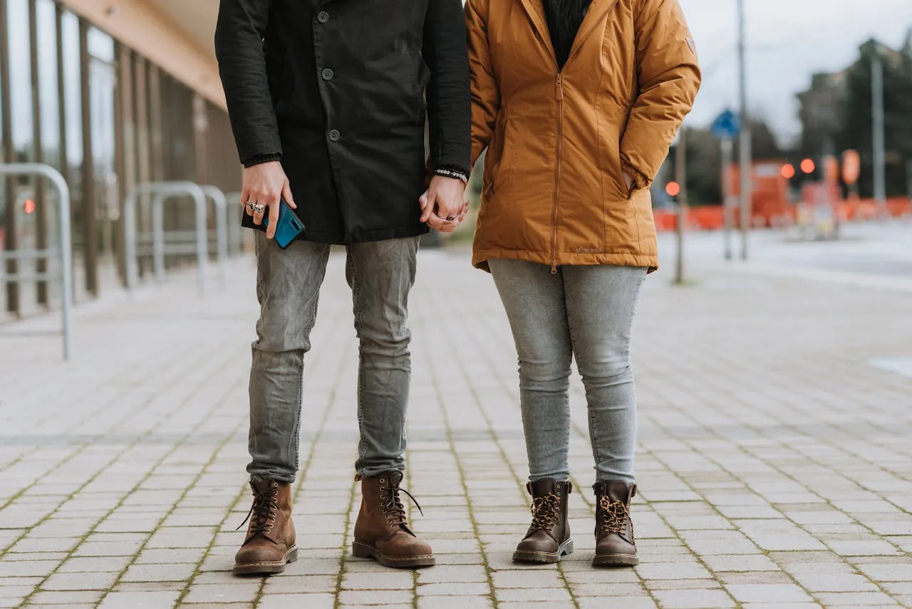 Couple standing on a paved sidewalk holding hands, wearing boots and jackets.