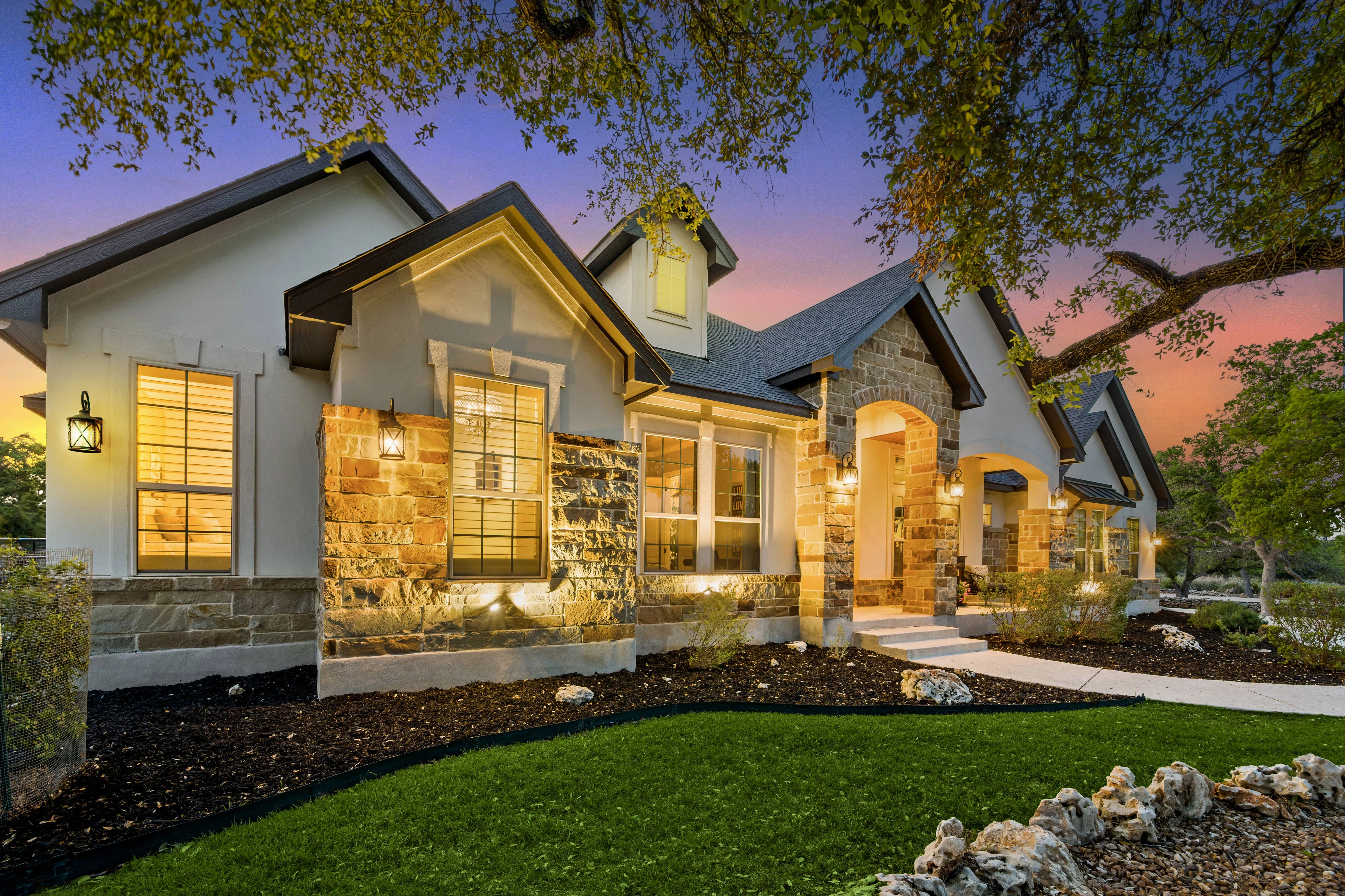 Illuminated suburban house exterior with stone and white walls at sunset, surrounded by trees and green lawn.
