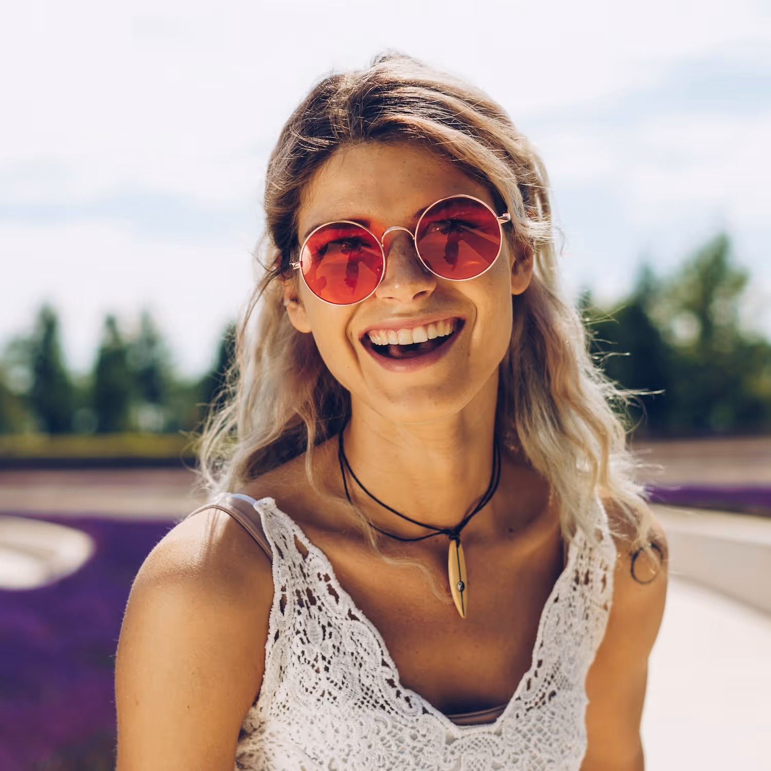 Smiling woman with wavy blonde hair wearing red round sunglasses, a white lace top, and a pendant necklace outdoors.