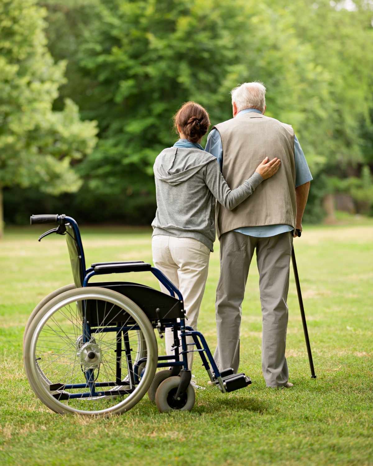 Woman supporting elderly man with a cane as they walk away from an empty wheelchair on grass.